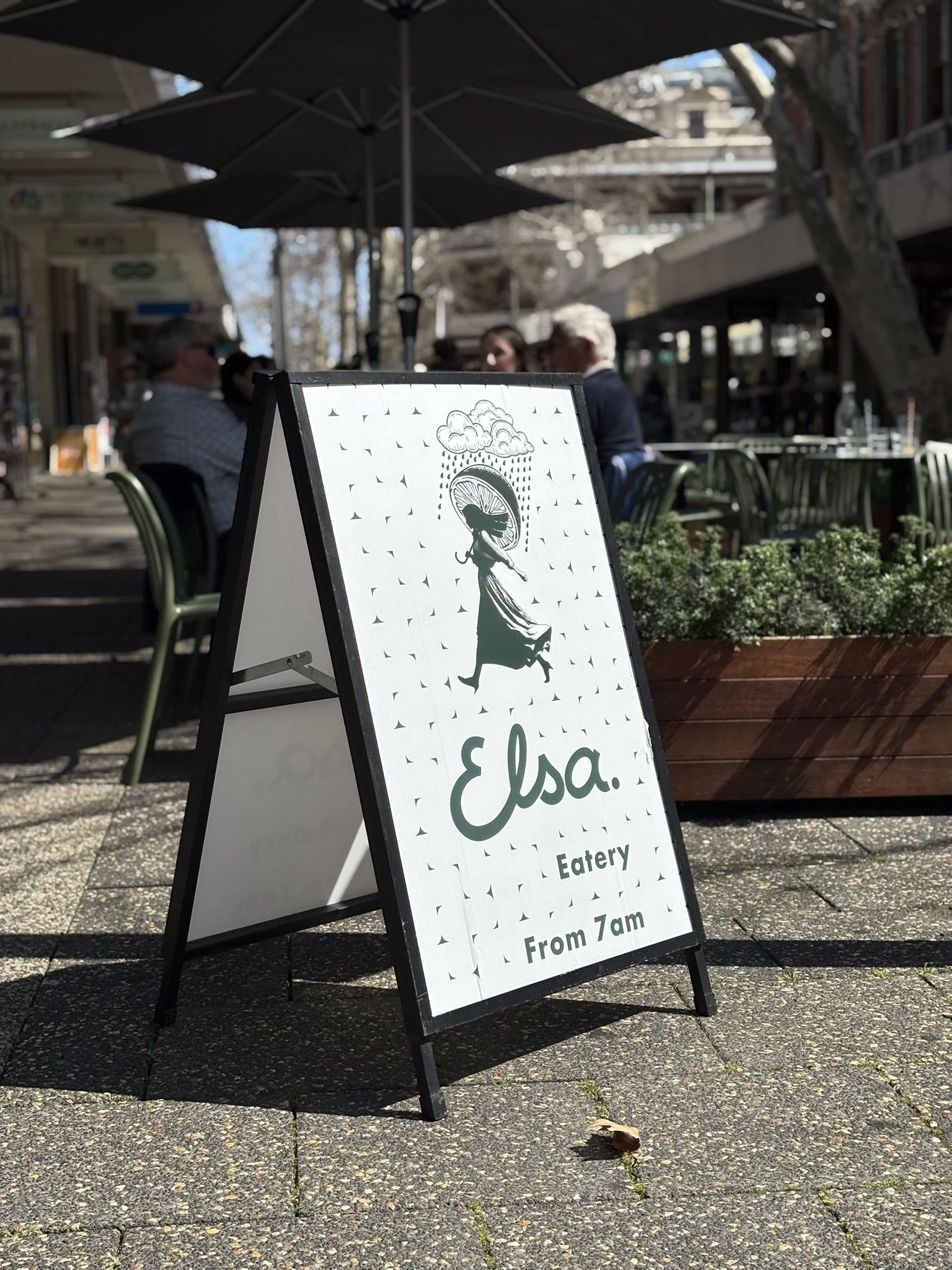 Aboard a sidewalk, a white and black sandwich board sign advertises Elsa Eatery, open from 7 am. The sign features a silhouette of a woman holding an umbrella and wearing a hat, with rain clouds above. In the background, people sit under umbrellas at outdoor tables.