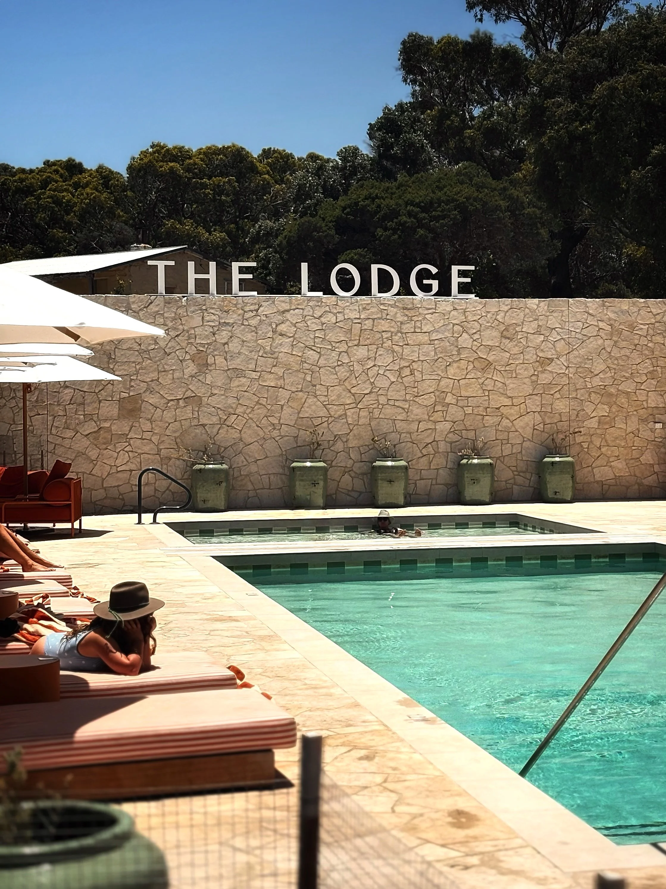 People relaxing by a swimming pool at a resort called The Lodge, with a stone wall, large planters, and trees in the background.
