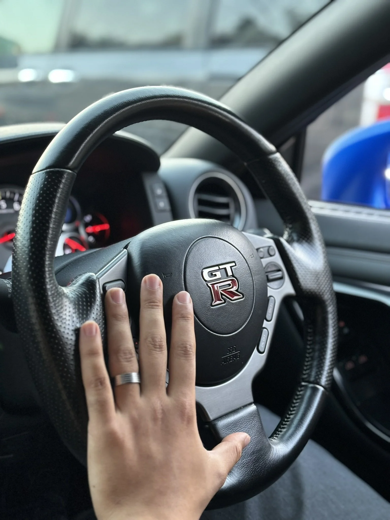 A person’s hand with a wedding ring on the ring finger placed on a car's steering wheel with GT R logo, inside a vehicle.