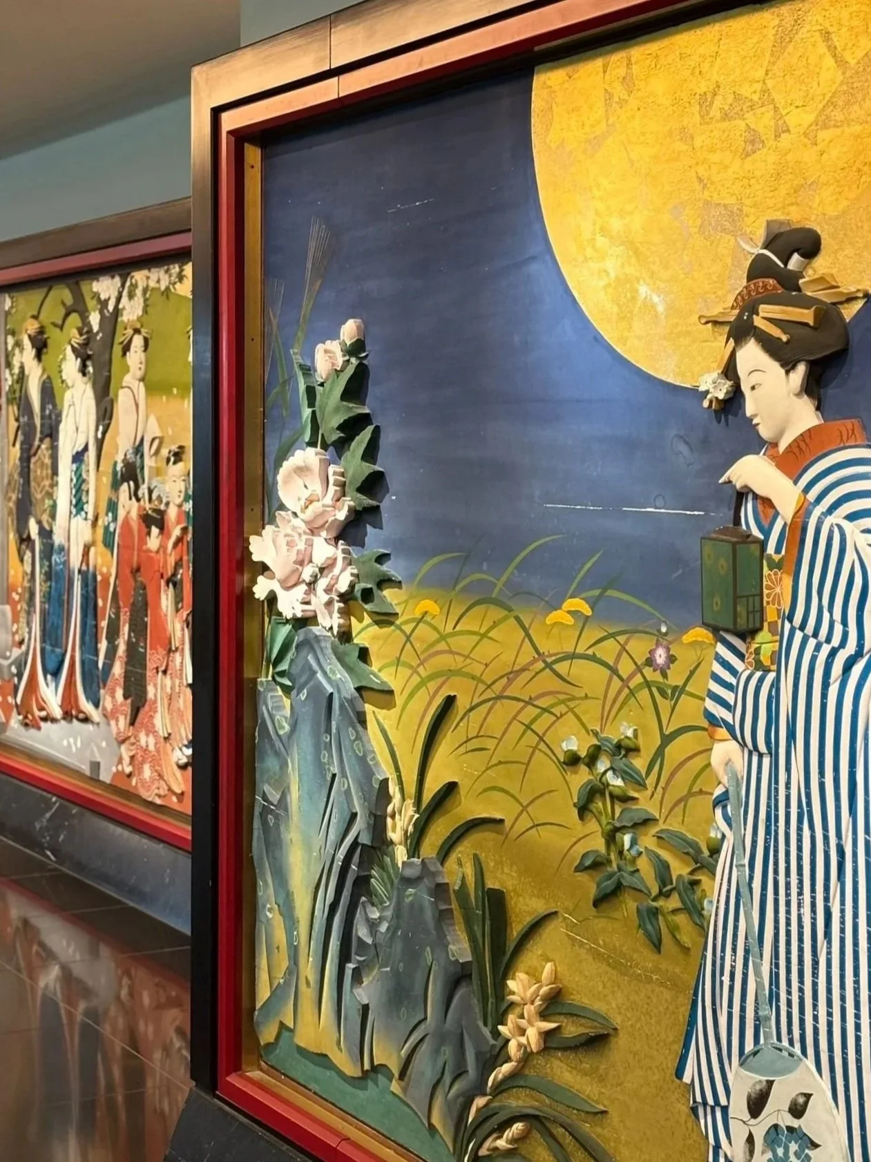 Japanese paper art display featuring a woman in traditional kimono holding a fan, with a large yellow moon in the background, flowers, and plants.