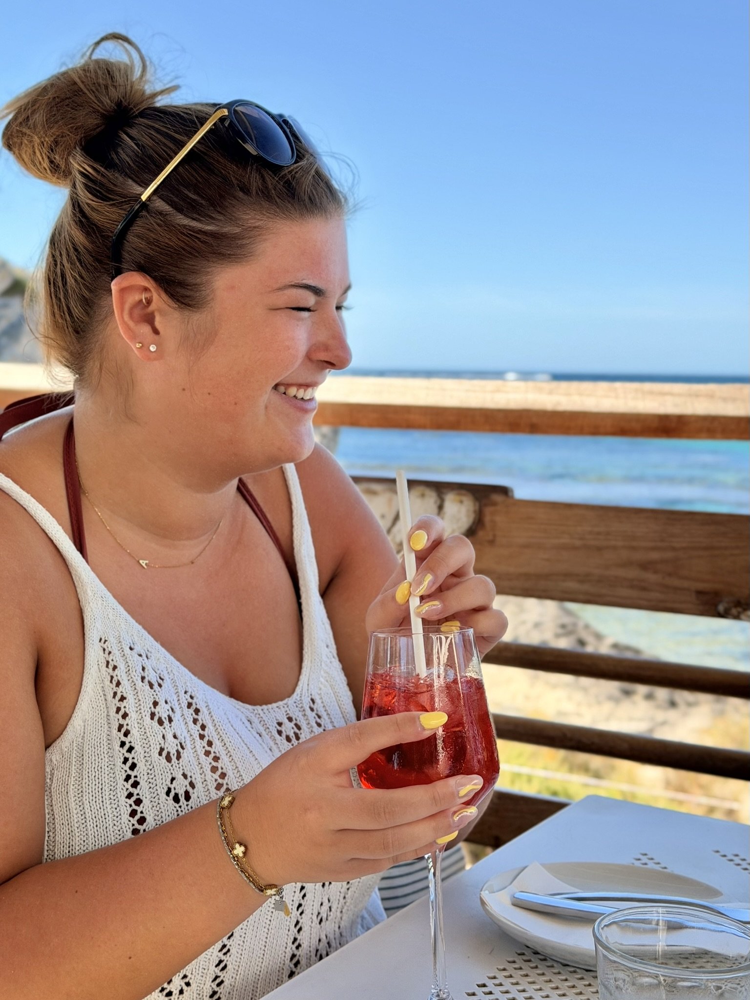A woman with sunglasses on her head is sitting outdoors at a restaurant, smiling and holding a glass of red beverage with ice and a straw, with a beach and ocean in the background.