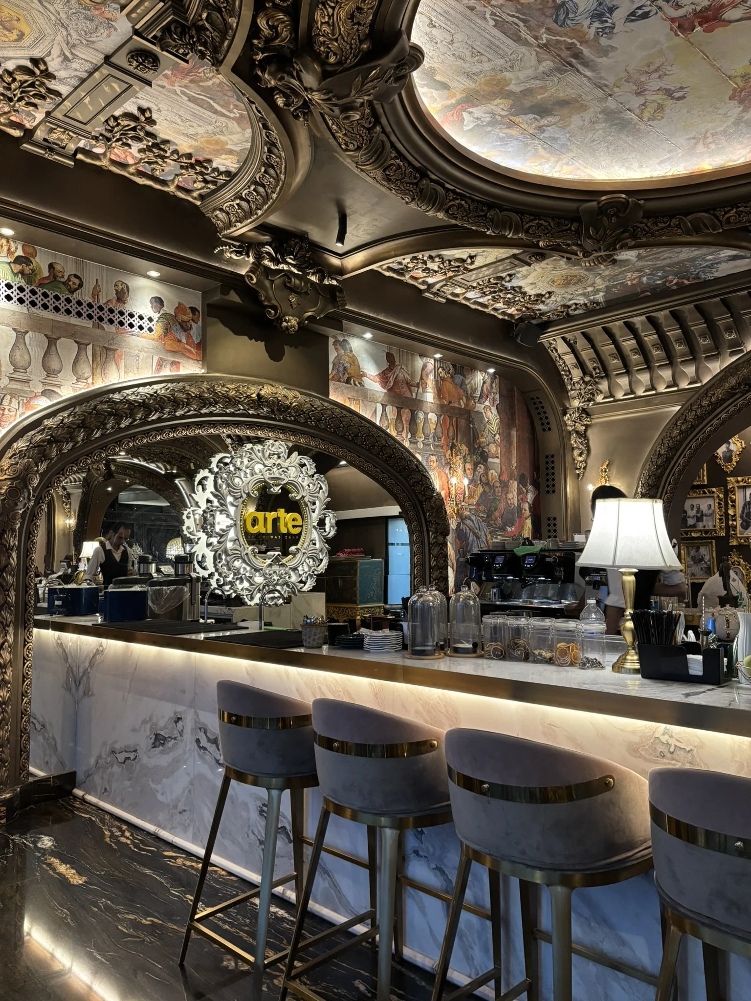 Elegant bar with marble counter and ornate decorative arch, featuring a sign that says 'arte' with barstools, lamps, and bar equipment, set in a richly decorated room with detailed ceiling murals and artwork.