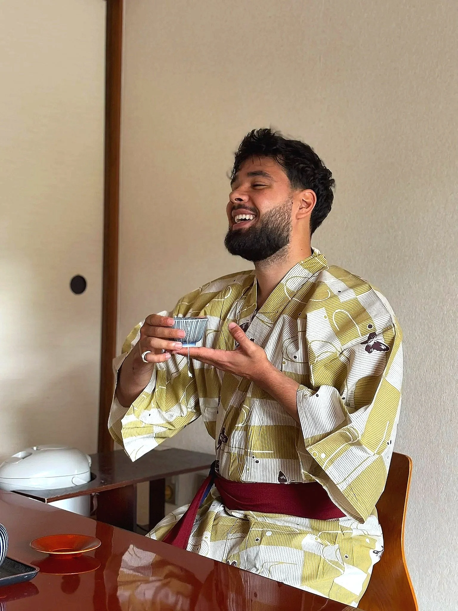 A man with a beard and short dark hair wearing a yellow and white checkered kimono with butterfly patterns, holding a small bowl, smiling with eyes closed, sitting at a dining table indoors.