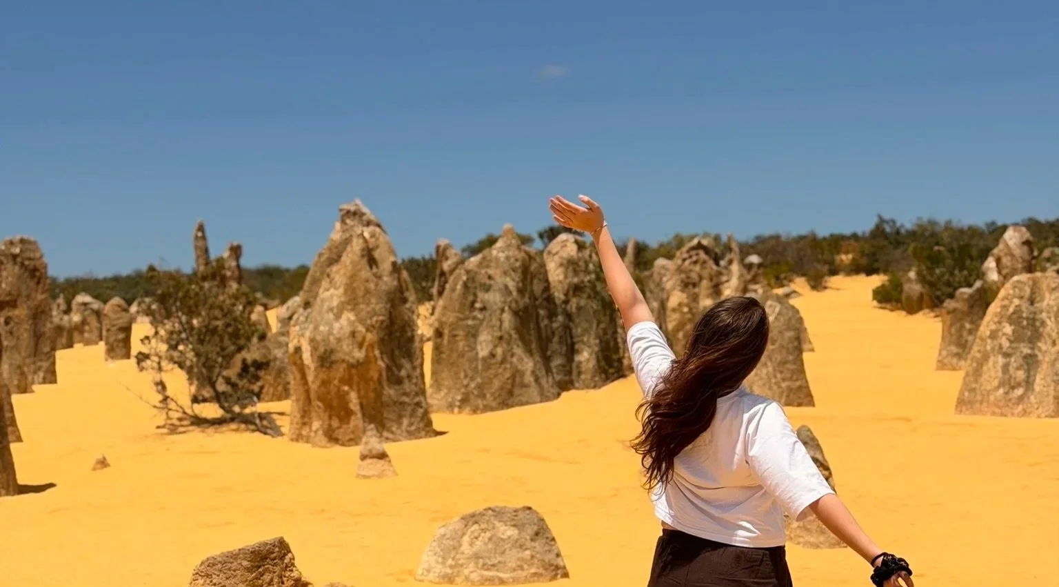 A woman with long brown hair wearing a white shirt and black shorts standing in a desert with yellow sand and large rock formations, raising her right arm towards the sky against a clear blue sky.