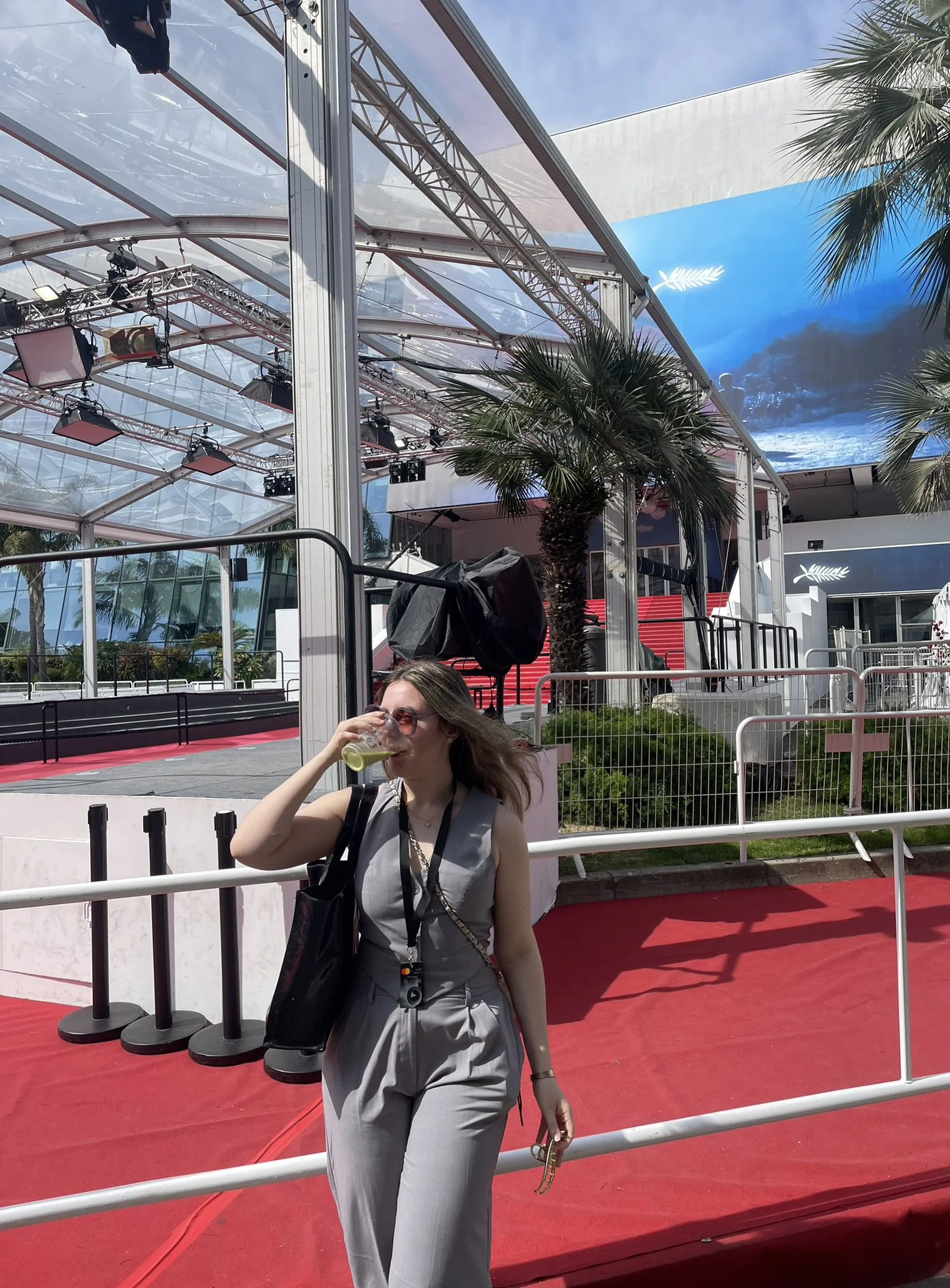 A woman in a gray jumpsuit drinking a yellow beverage at an outdoor event space with palm trees, a stage, and a large LED screen in the background.