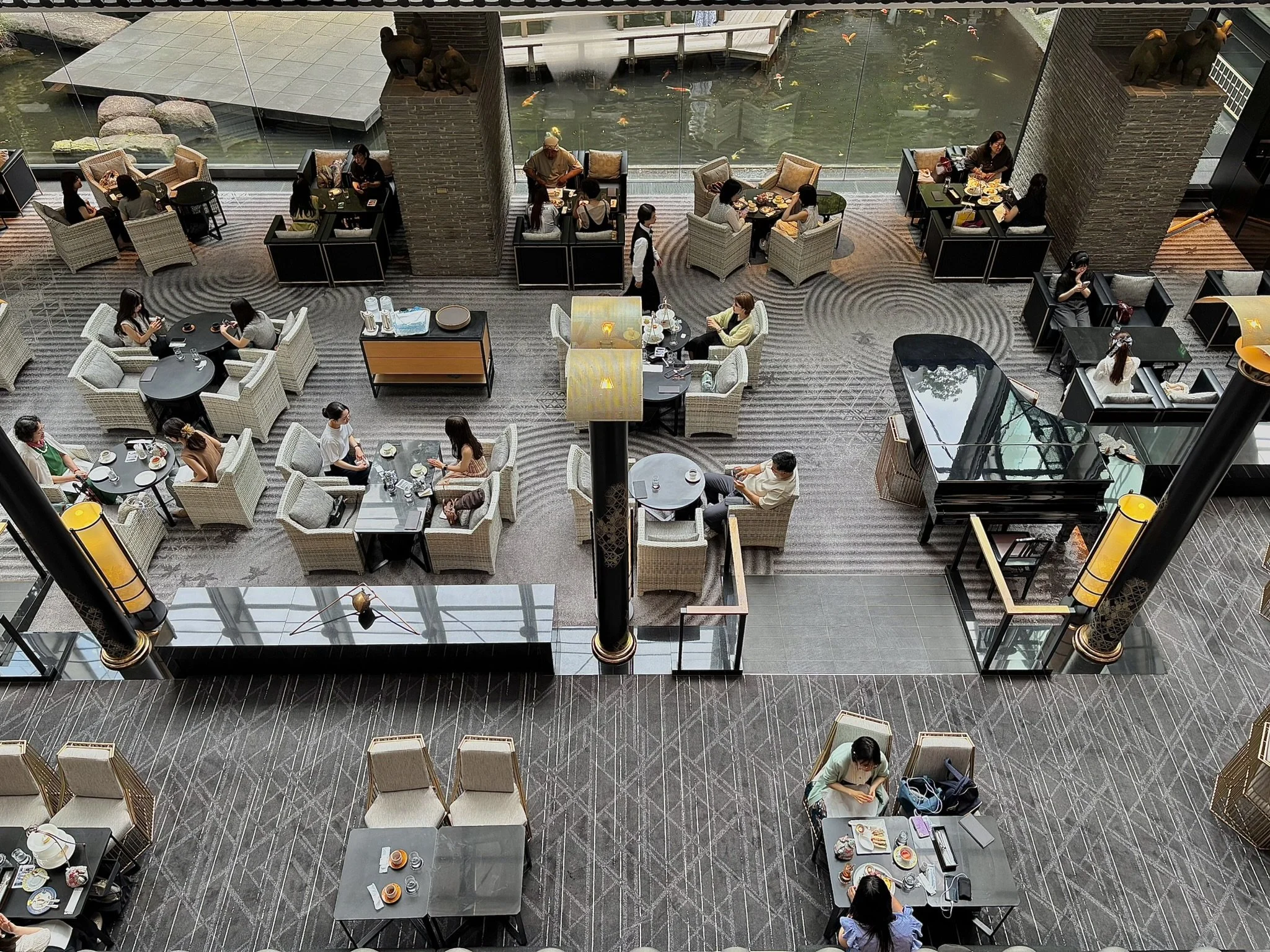 Interior view of a restaurant with various tables occupied by diners. Large glass windows overlooking a water pond with koi fish. Elegant decor includes a black grand piano and traditional Chinese lanterns.