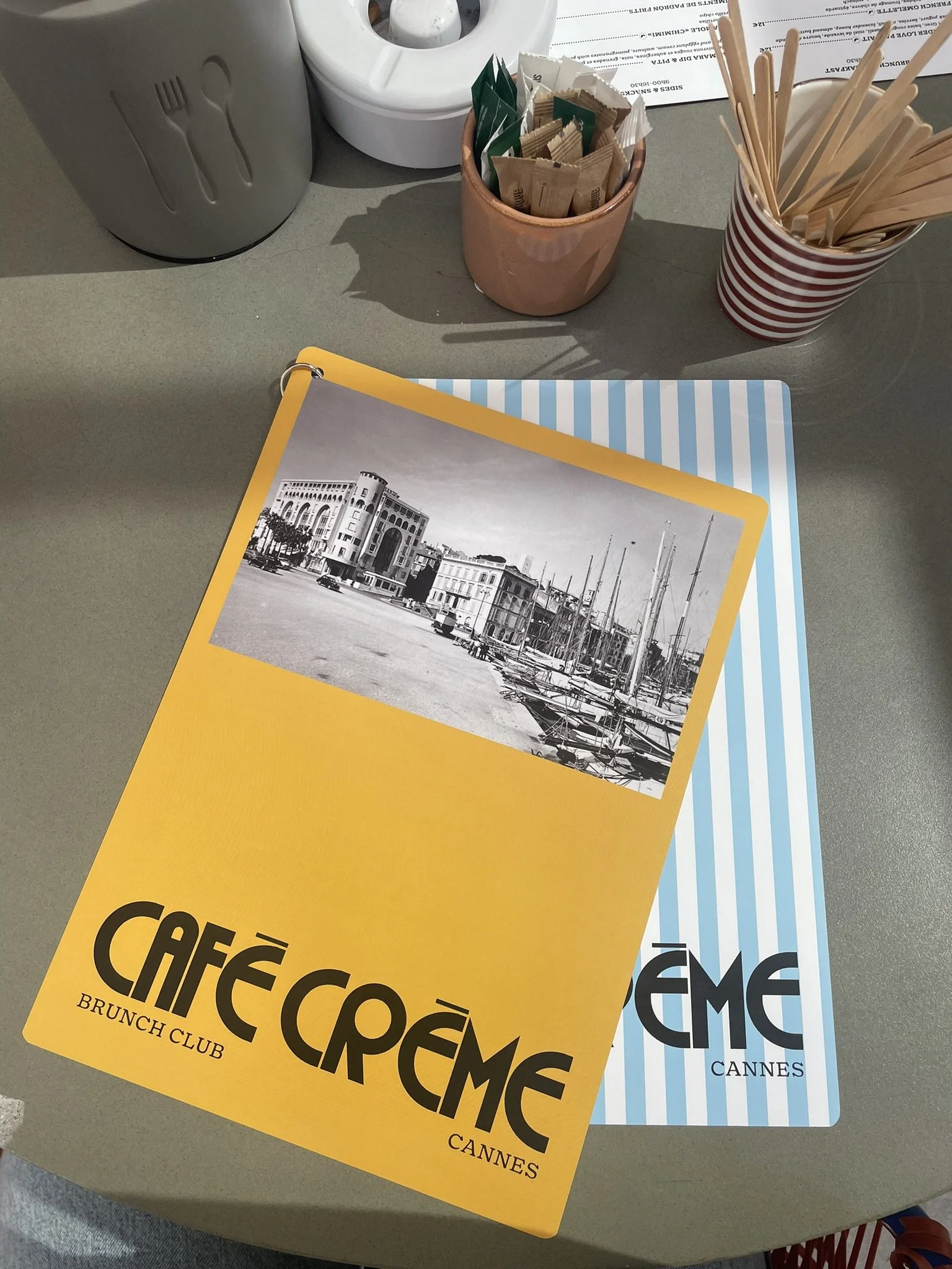 Close-up of two menus for Café Càréme in Cannes, placed on a gray table with various utensils and condiments at a cafe.