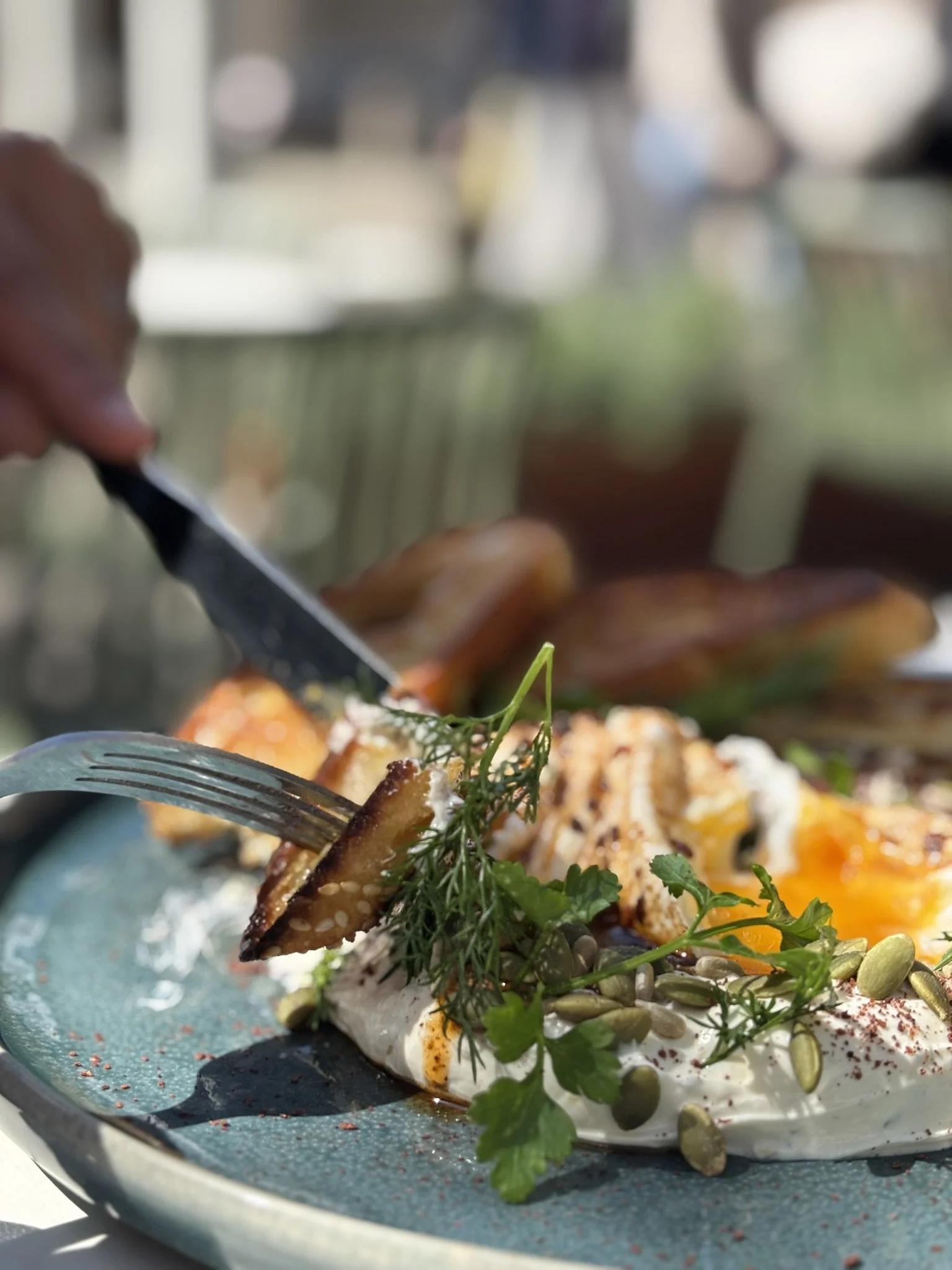 Close-up of a plate of food with eggs, herbs, and seeds, with a hand using a fork and knife to eat a piece of toasted bread.