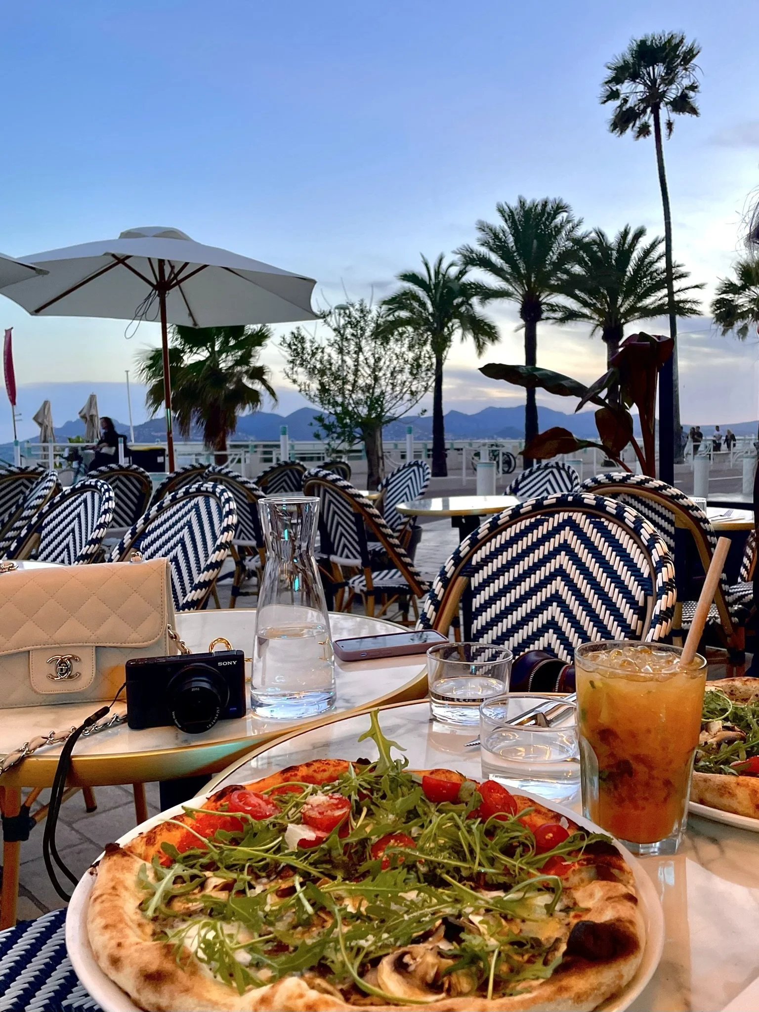 Outdoor restaurant scene with a table featuring a pizza with arugula and cherry tomatoes, a glass of iced tea, a carafe of water, glasses, and a white Chanel purse. Background includes striped chairs, palm trees, a mountain range, and a sunset sky.