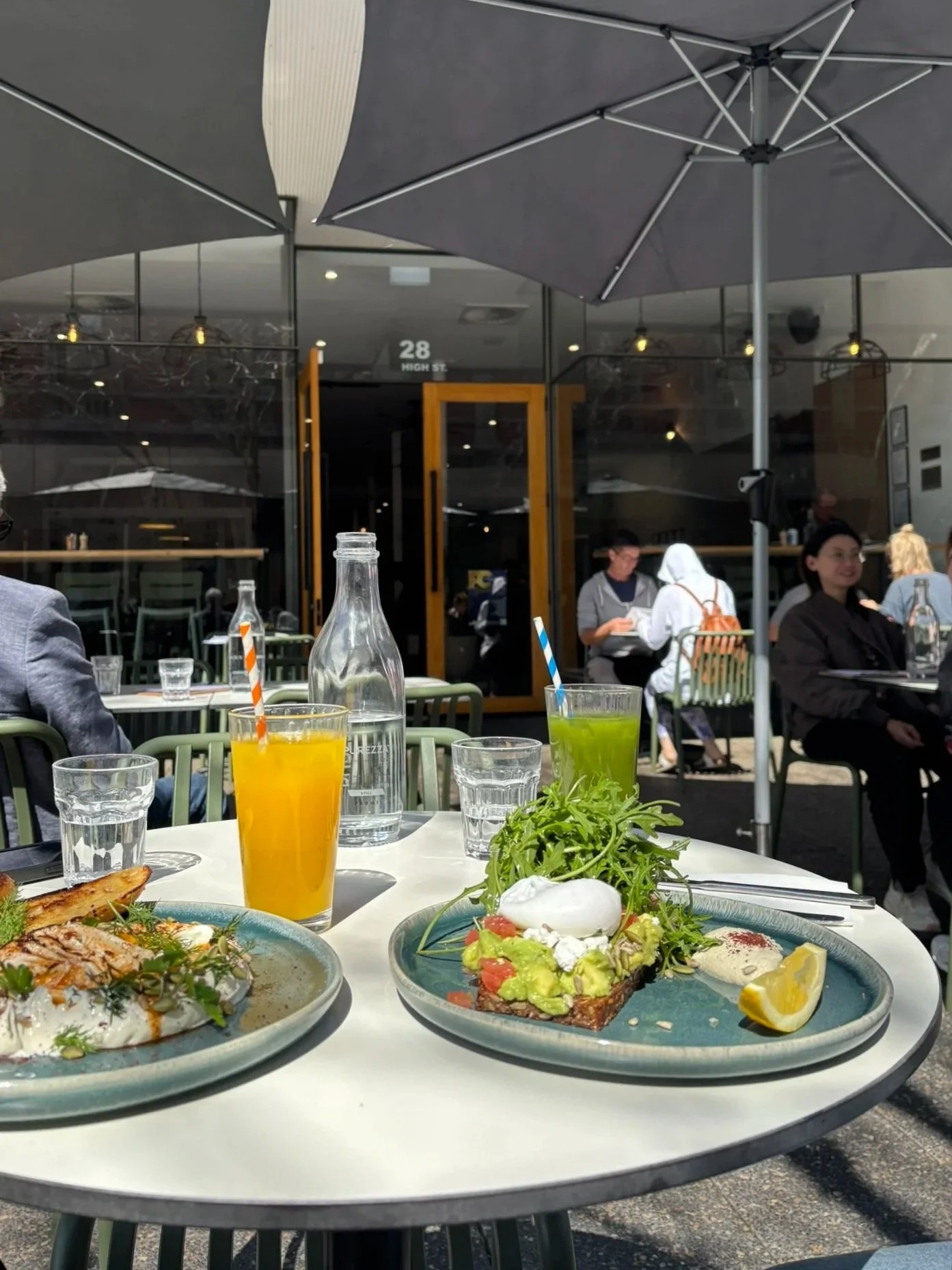 Outdoor restaurant patio with a table of food and drinks, including toast with toppings, a green juice, an orange juice, a glass of water, and a water bottle. Several people are seated in the background, and large umbrellas shade the table.