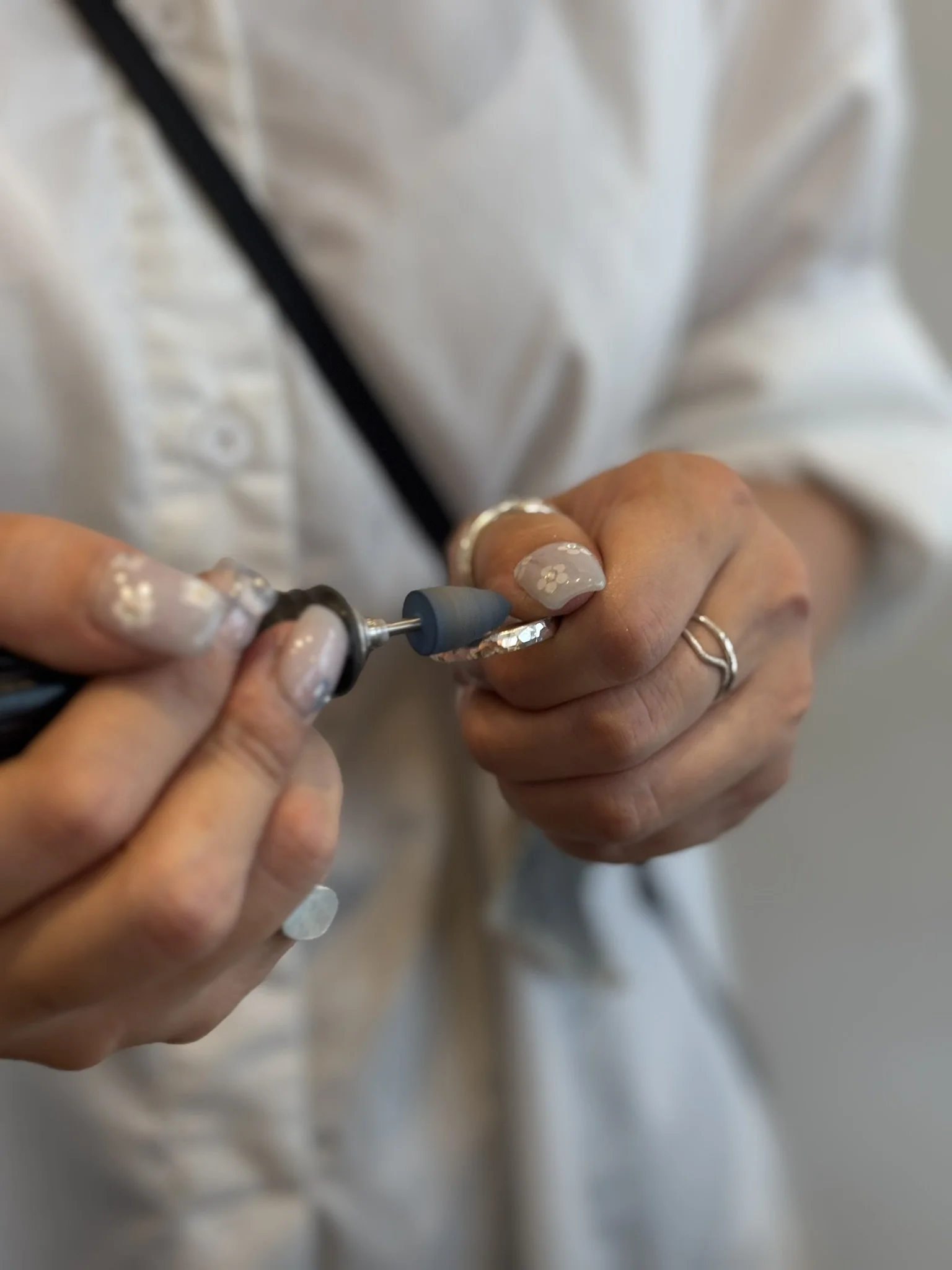 Hands holding a nail drill, preparing to file or shape a nail, with a background of a person wearing a white coat.