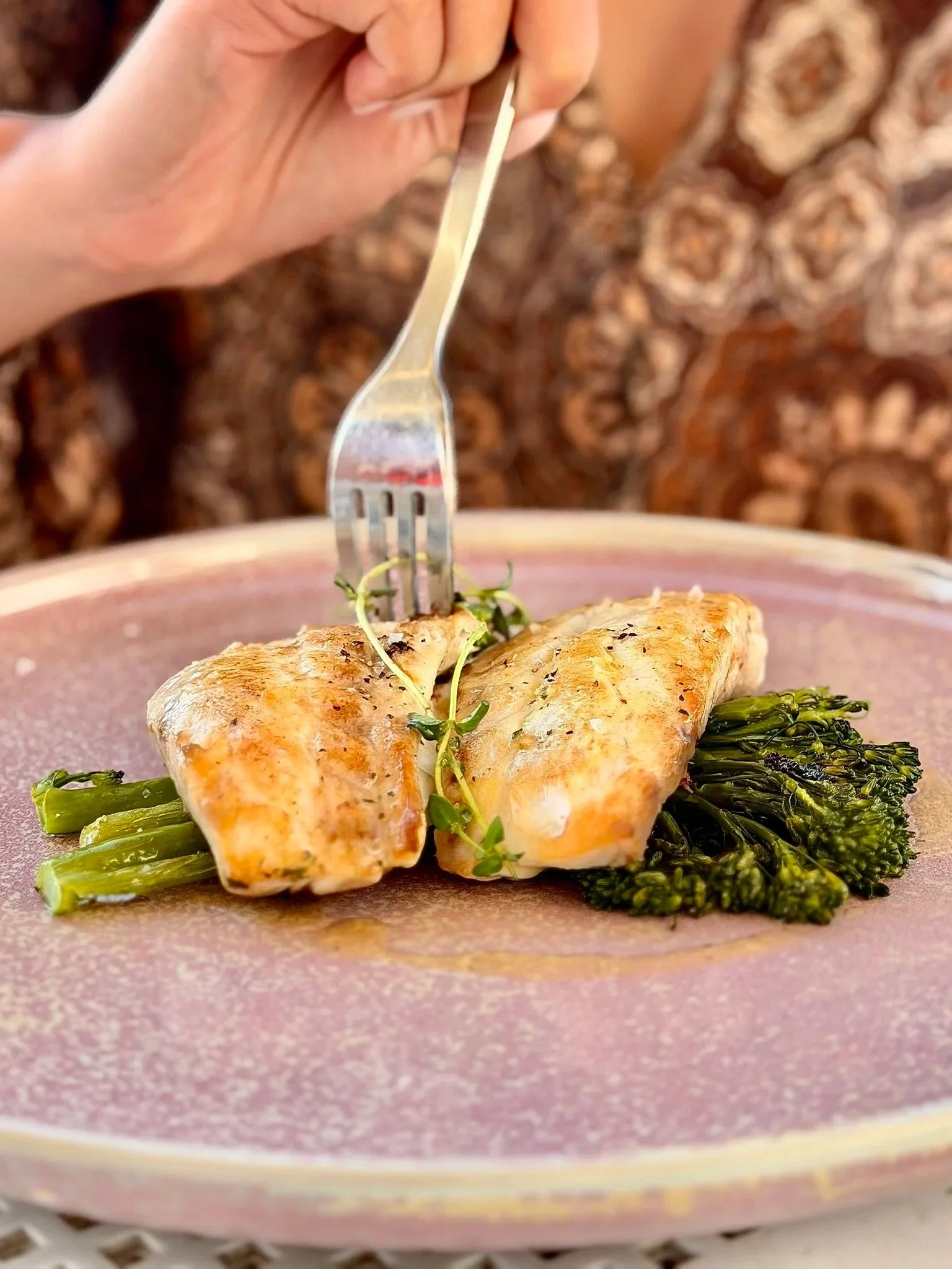 Close-up of grilled chicken breast with asparagus and microgreens on a pink plate, a hand holding a fork above the food.