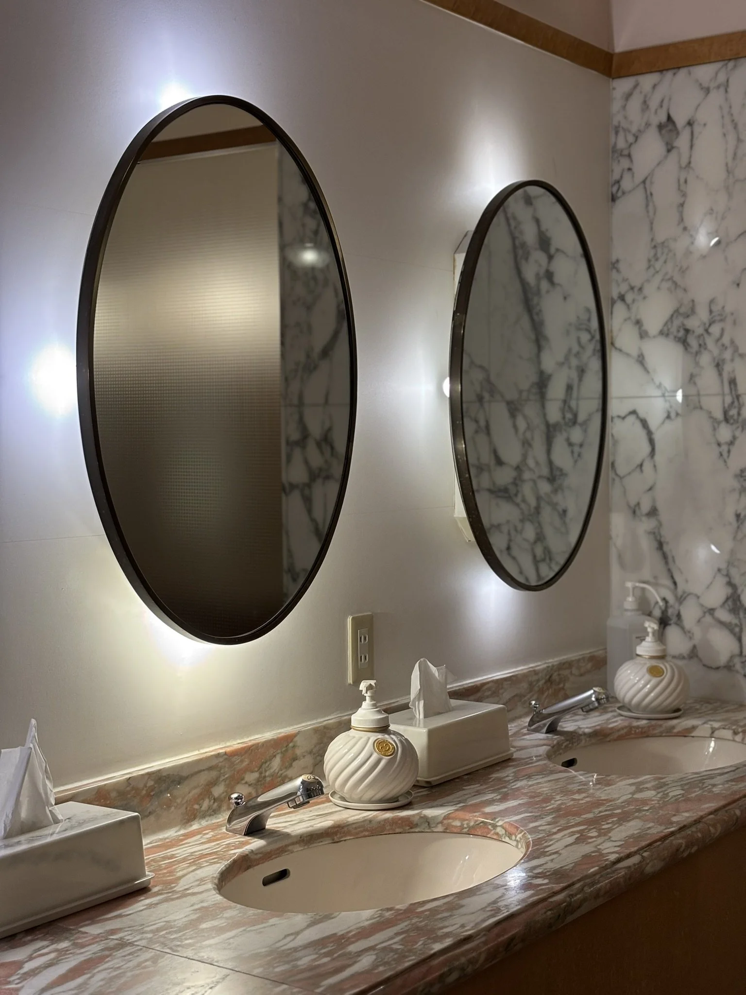 A bathroom vanity with two sinks, marble countertops, two oval mirrors with backlit lighting, tissue box, liquid soap dispensers, and a marble wall.