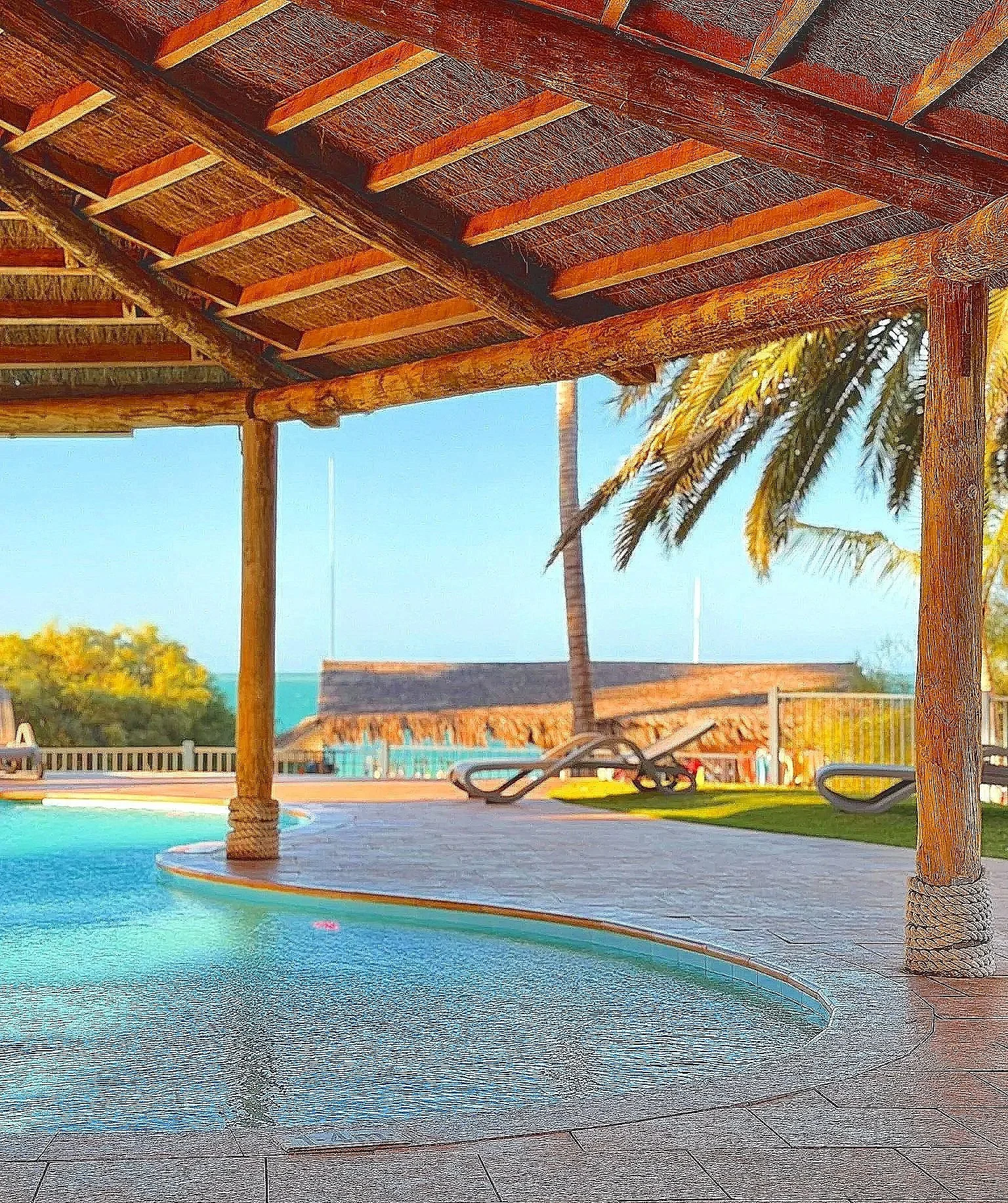 Swimming pool under a wooden pergola with lounge chairs, palm trees, and a thatched roof building in the background, sunny sky.