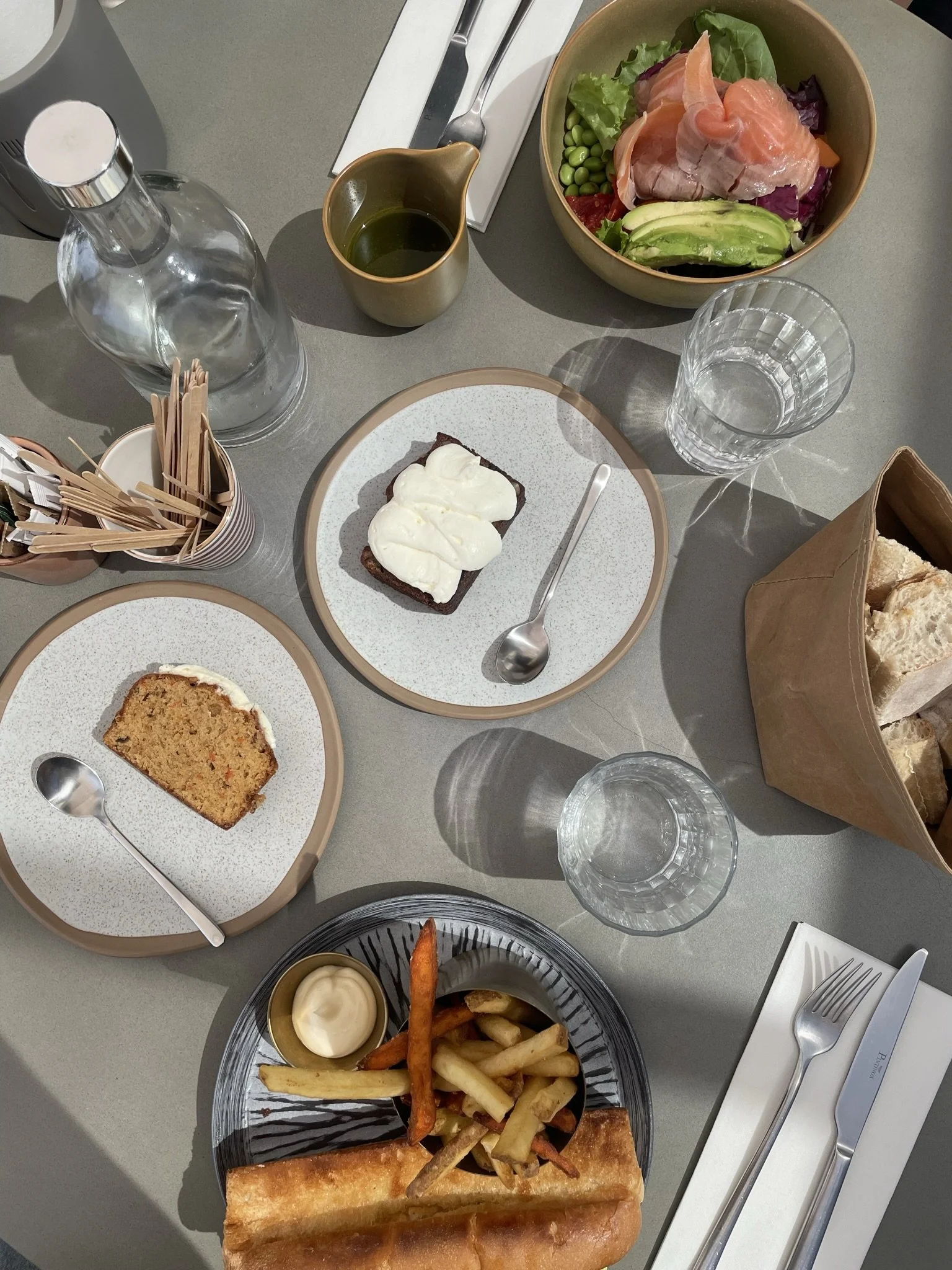 A table set for a meal with various dishes including a bowl of salad with meats and vegetables, a plate of carrot and potato fries with mayonnaise, slices of bread in a paper bag, a slice of carrot cake and a dessert with whipped cream, a glass bottle of water, glasses of water, and condiments, with cutlery and napkins.
