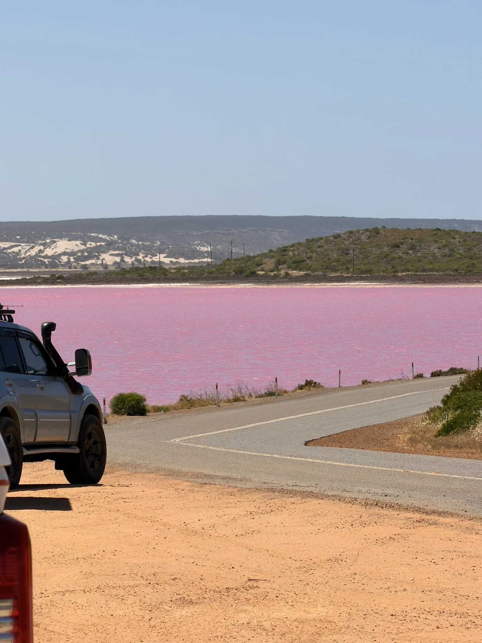 Pink salt lake with hills in the background, part of a parking area with cars visible in the foreground.