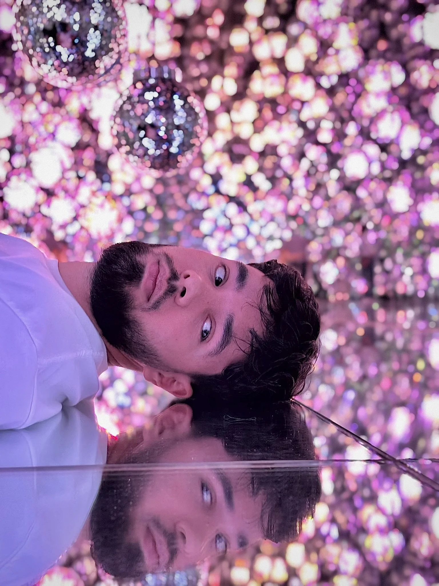 A man with dark hair and a beard lying down with a reflective surface beneath him. He is wearing a white shirt and looking at the camera with one eye winking. The background features a vibrant, colorful display of pink, purple, and white lights and decorations, with a disco ball visible at the top.