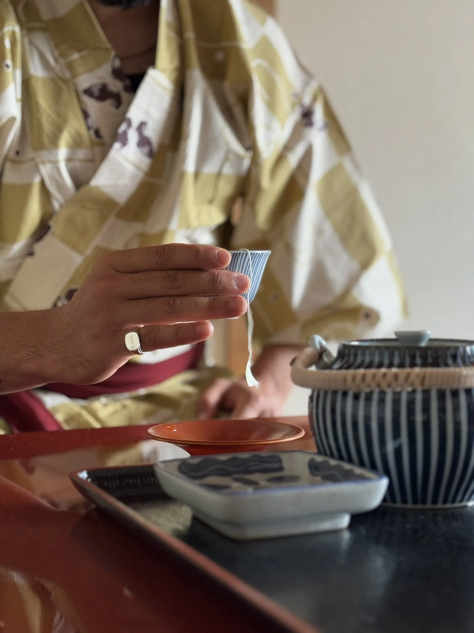 A person in a yellow patterned shirt is holding a blue and white ceramic tea scoop over a traditional Japanese tea bowl, with various small ceramic dishes and a tea container on a tray.