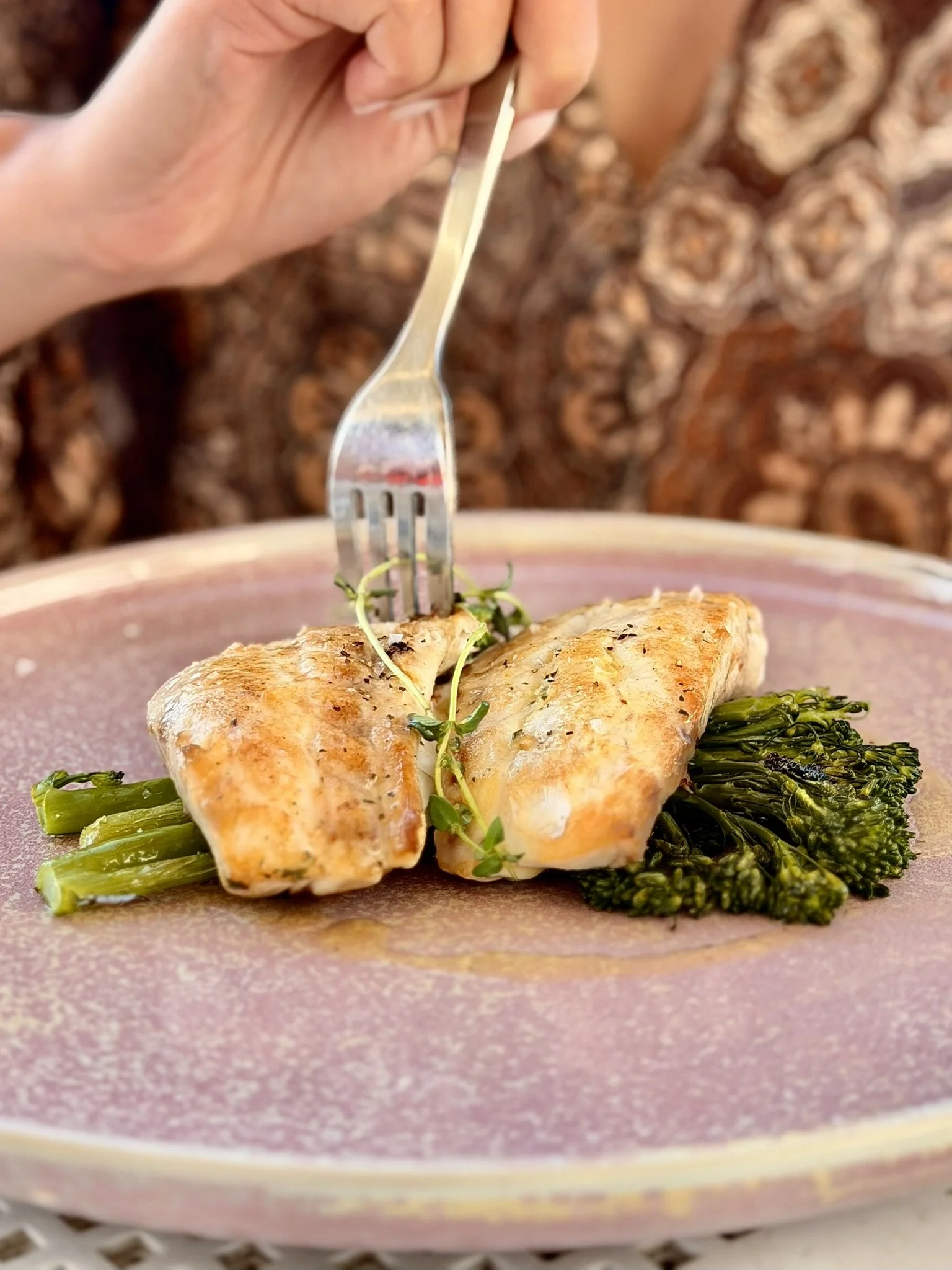 A fork piercing a cooked piece of chicken served with green vegetables on a pink plate.