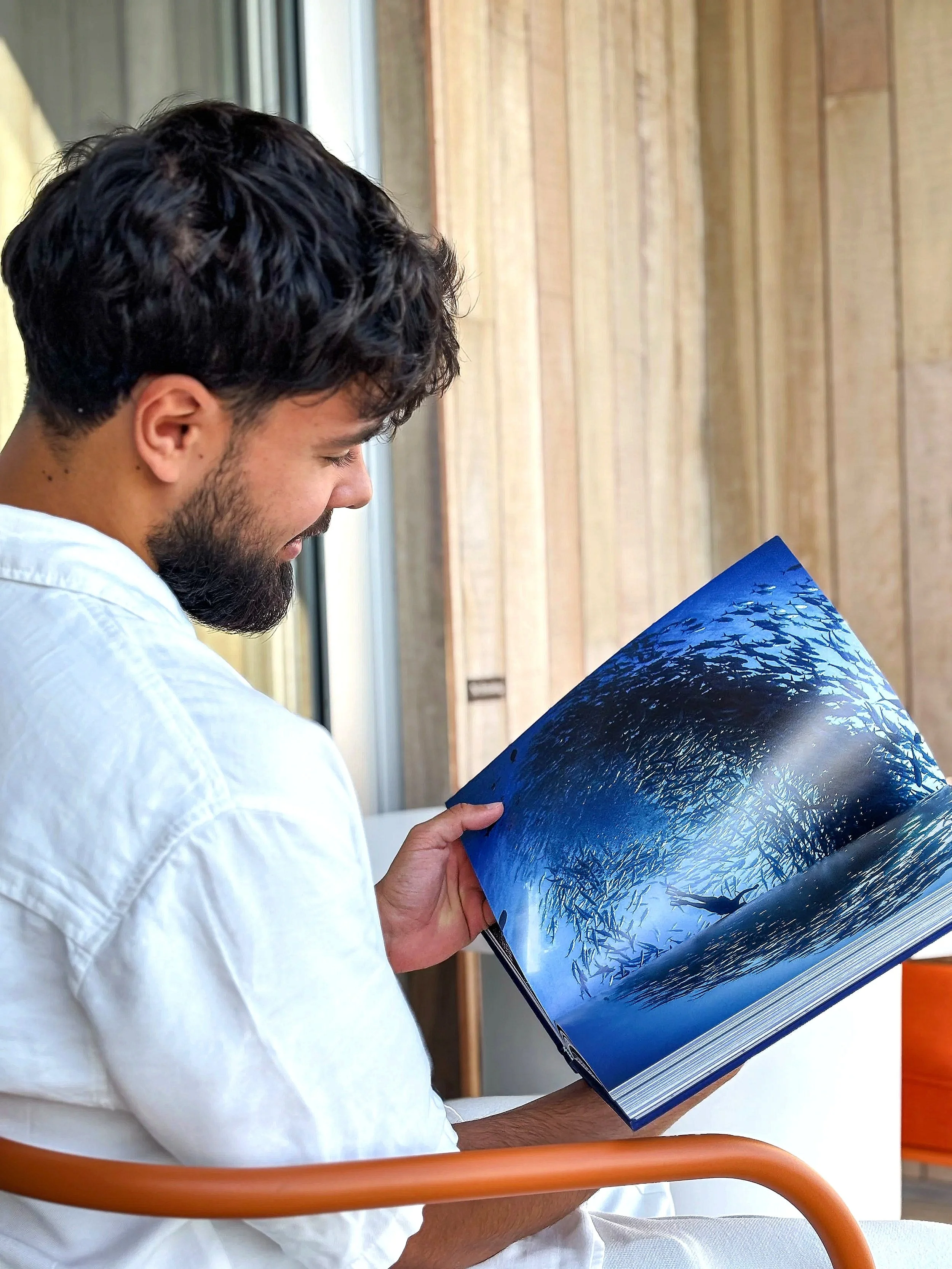 Man with dark curly hair and beard sitting in a chair, reading a large book with a beach or lake scene on the pages, in a room with wooden paneling and a large window.
