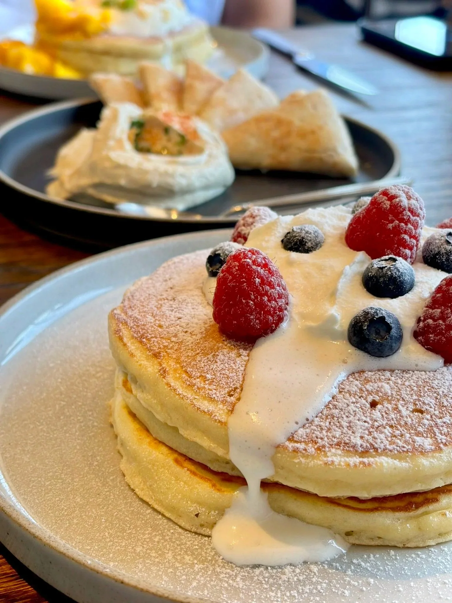Stack of three fluffy pancakes topped with whipped cream, fresh raspberries, blueberries, and dusted with powdered sugar.