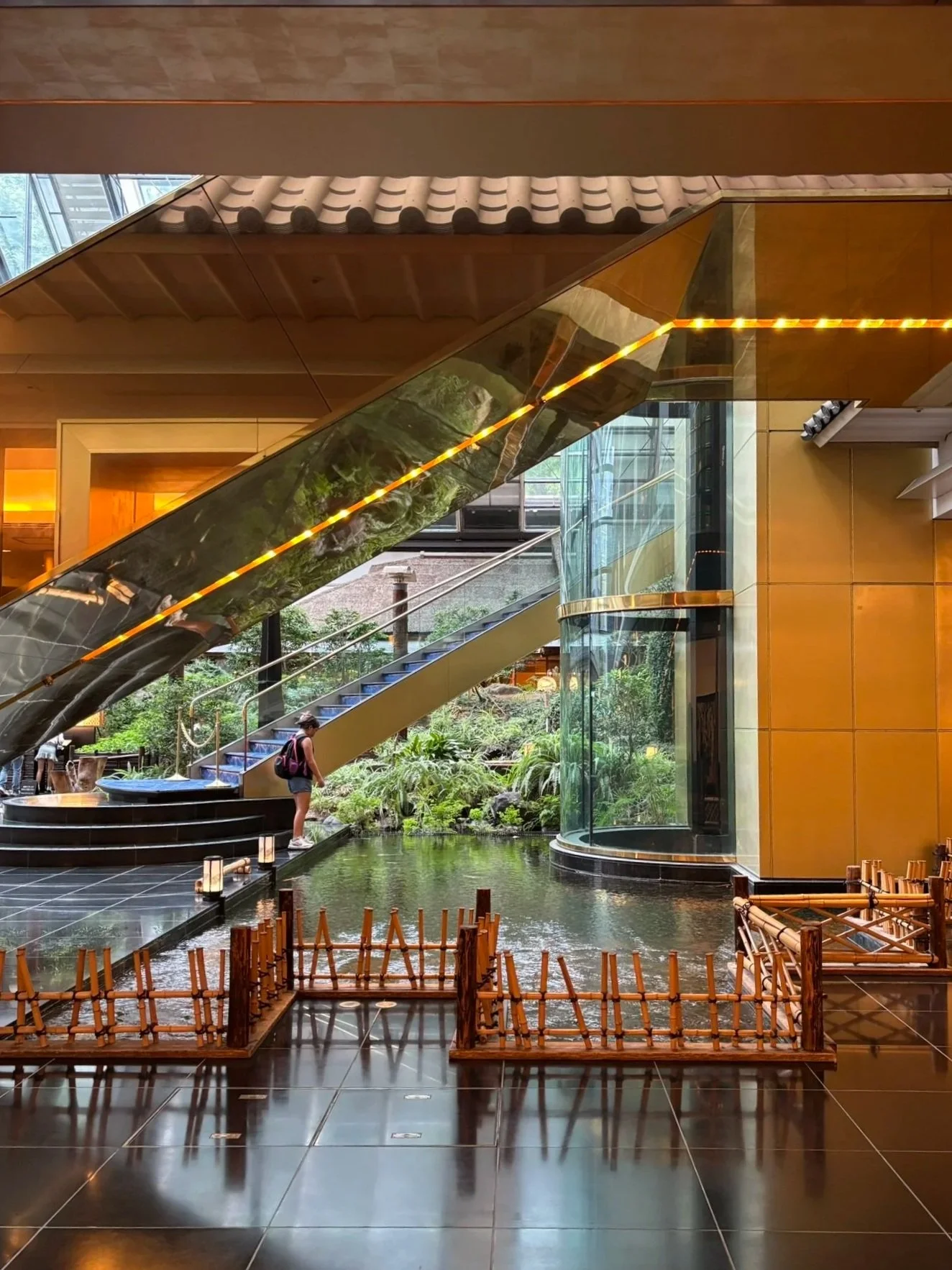 Interior view of a modern building with a water feature, a staircase, and lush greenery outside, featuring warm lighting and contemporary architectural design.