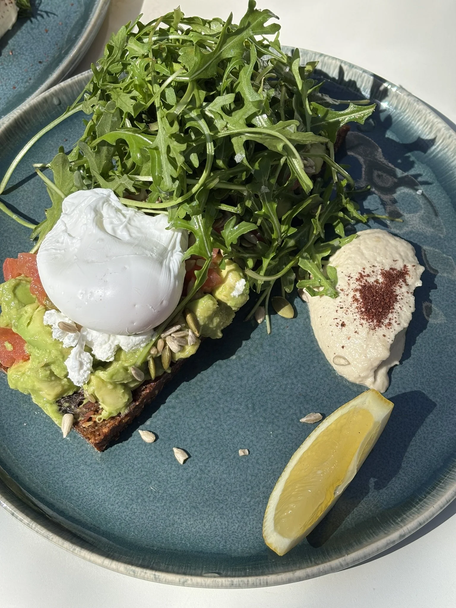 Open faced toast topped with guacamole, sour cream, and a poached egg, side of leafy greens, lemon wedge, and creamy dip with red seasoning.
