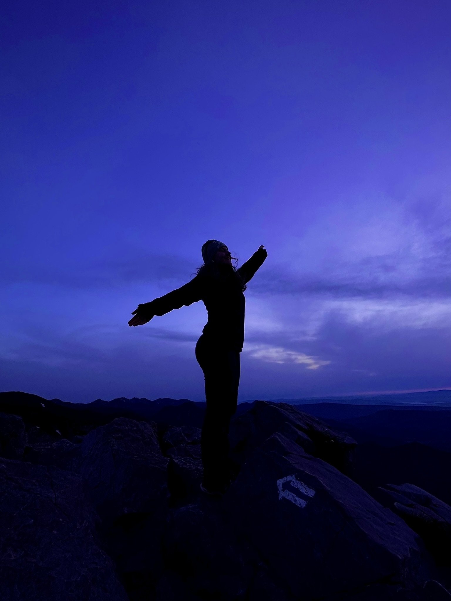 Silhouette of a person standing on rocks with arms outstretched, against a twilight sky with purple and blue hues, mountains in the distance.