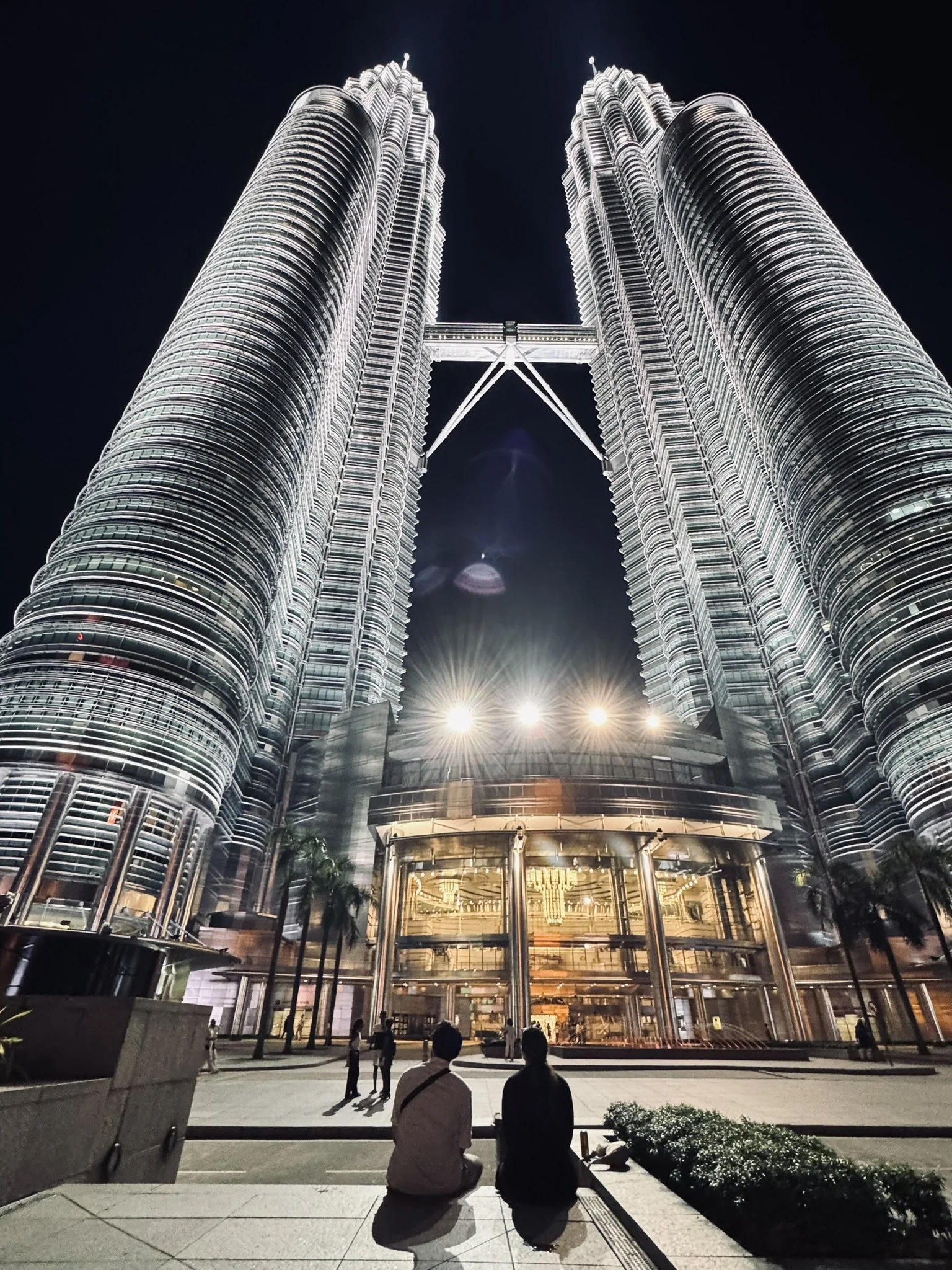 Night view of the Petronas Towers in Kuala Lumpur, Malaysia, with people sitting on the ground in the foreground.