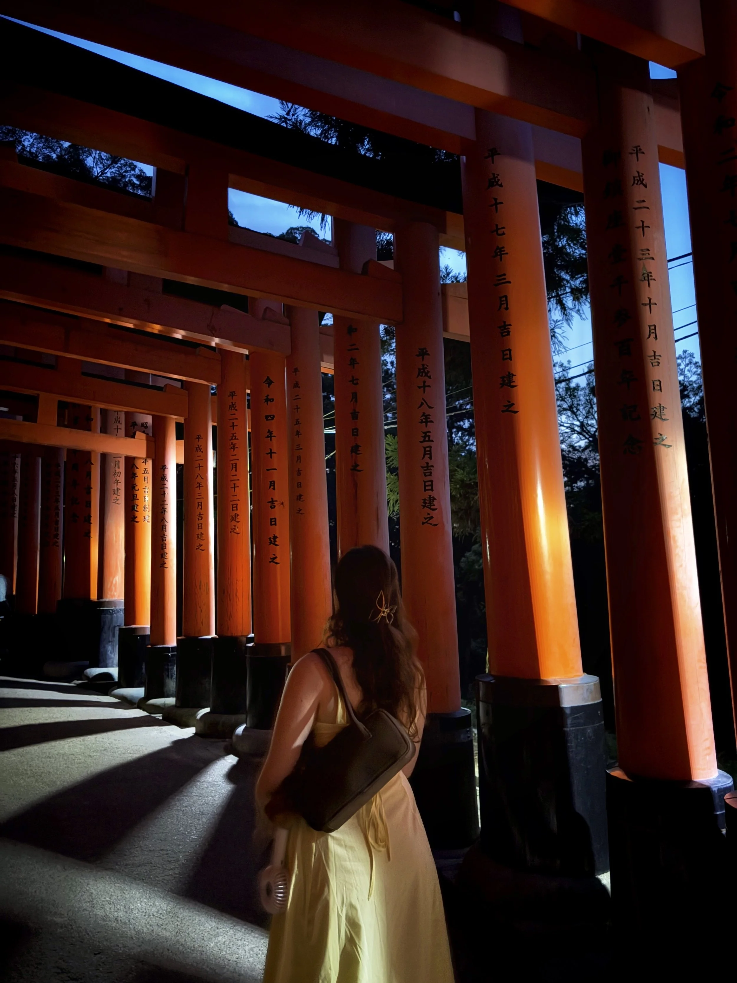 A woman in a yellow dress with a black shoulder bag walking near orange torii gates at dusk.