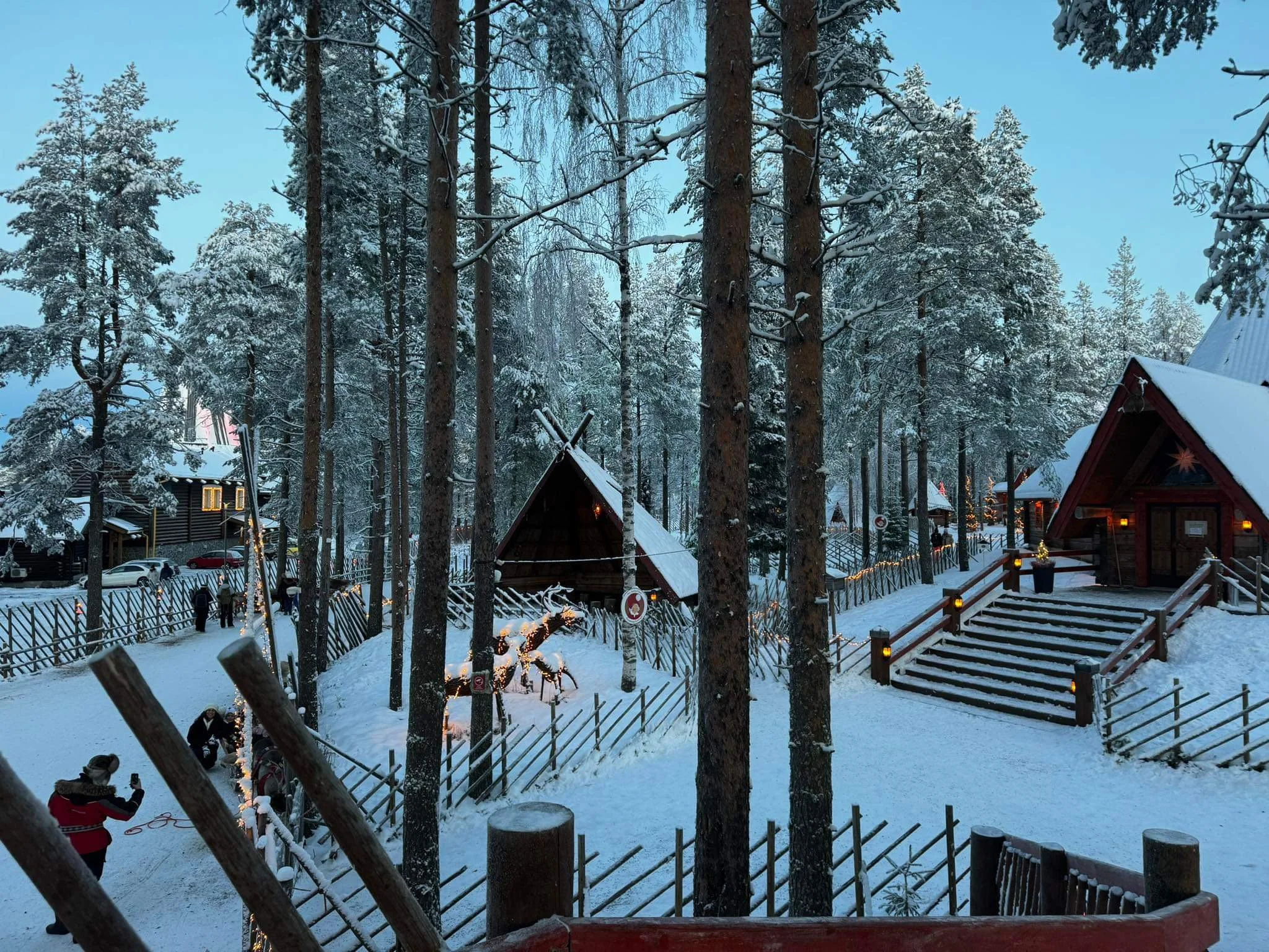 A snow-covered landscape in a forest with traditional wooden buildings and cabins, some decorated with string lights, and trees coated with snow.
