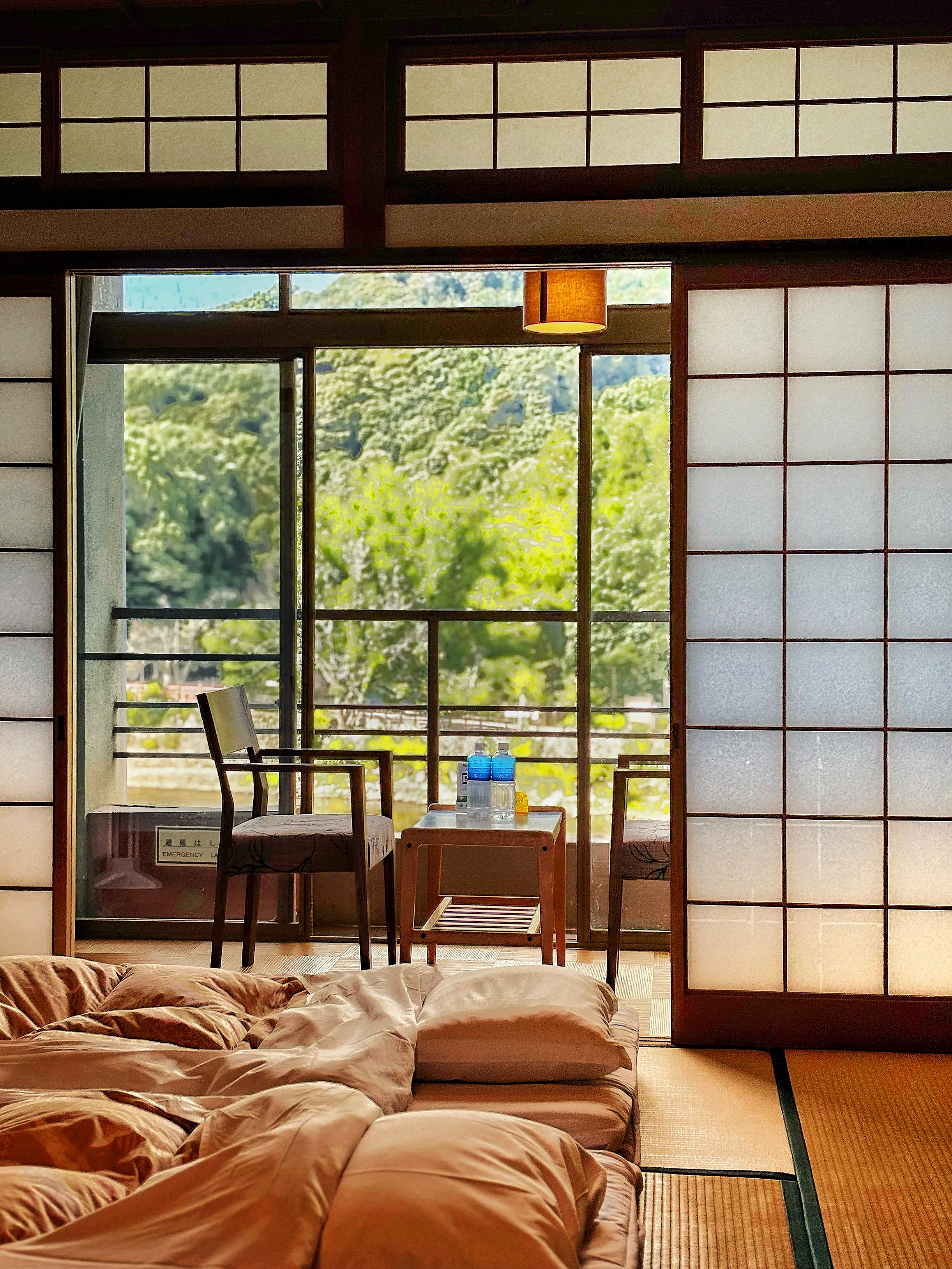 A traditional Japanese room with sliding shoji doors, tatami mats, and a view of a balcony with chairs and a table with water bottles, overlooking a lush green landscape.