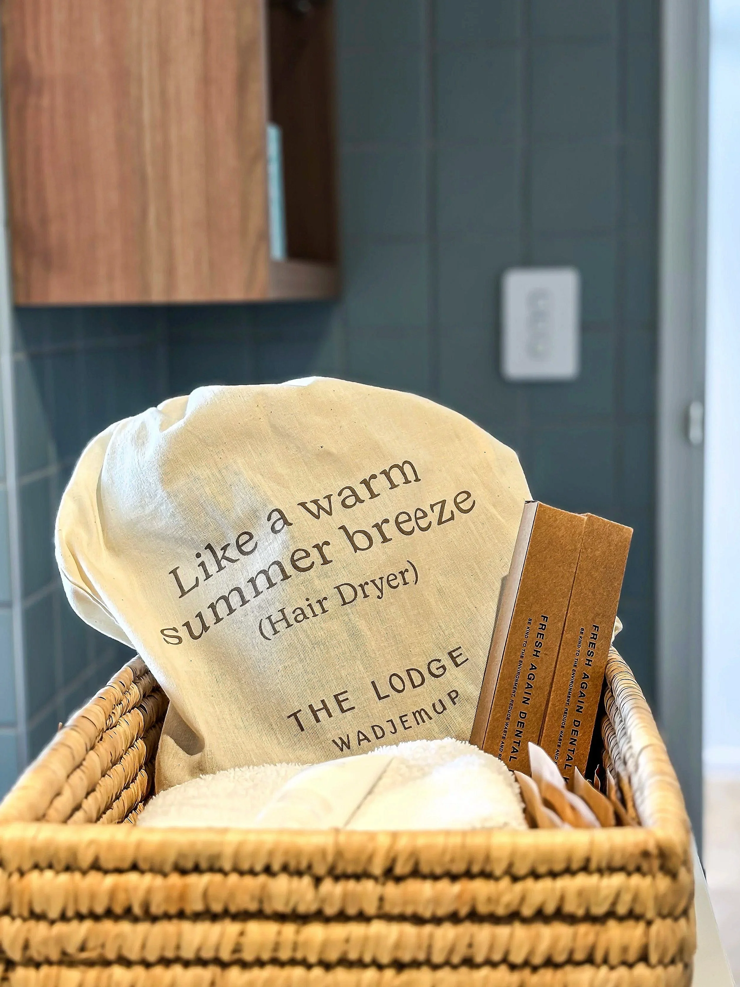 Wicker basket with a cloth bag that reads 'Like a warm summer breeze (Hair Dryer) THE LODGE WADJEMUP' and hair care promotional items inside.