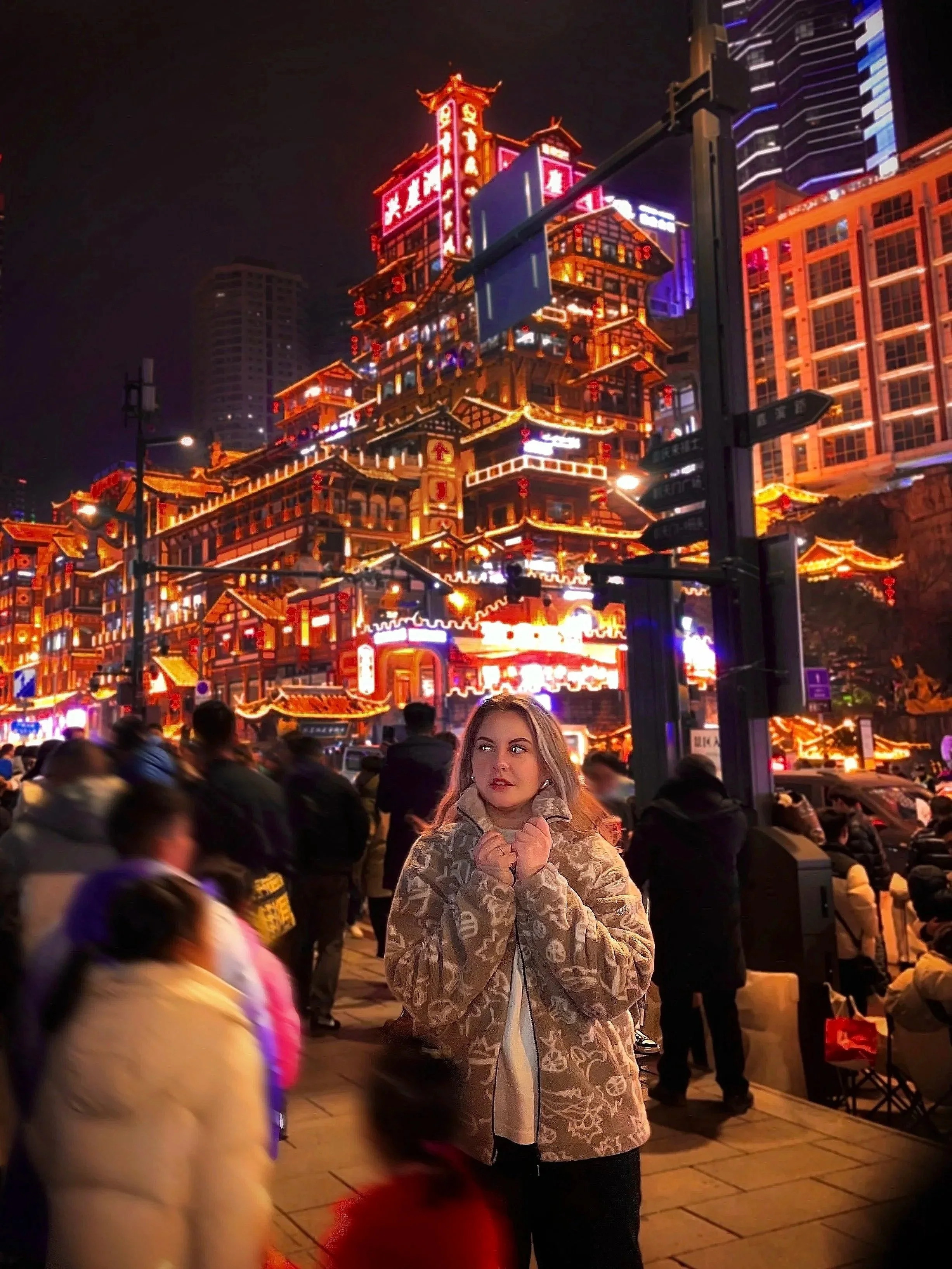 A young woman stands on a busy city street at night, wearing a patterned beige coat with her hands near her face. Behind her, tall buildings with vibrant neon lights and illuminated signs create a colorful backdrop. Several people walk or stand around, some blurred, indicating movement.