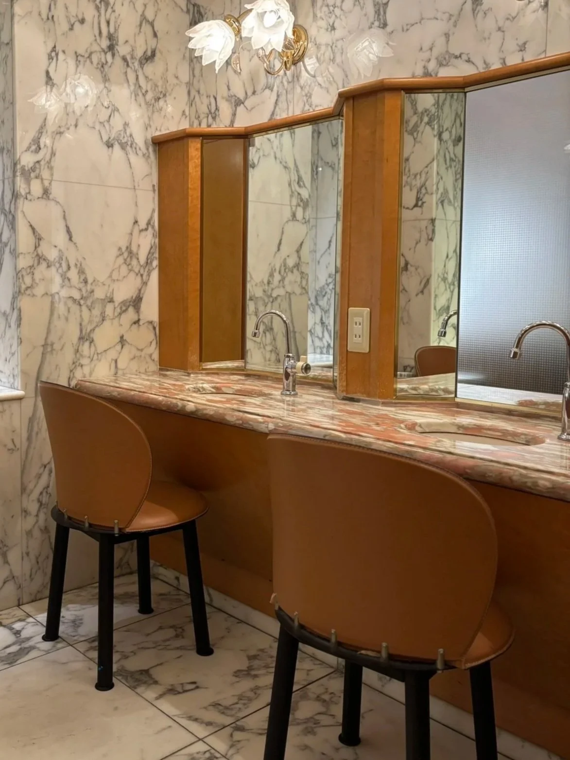 A vintage bathroom vanity with a marble countertop, two sinks, mirrors, and mid-century chairs in a marble-walled room.