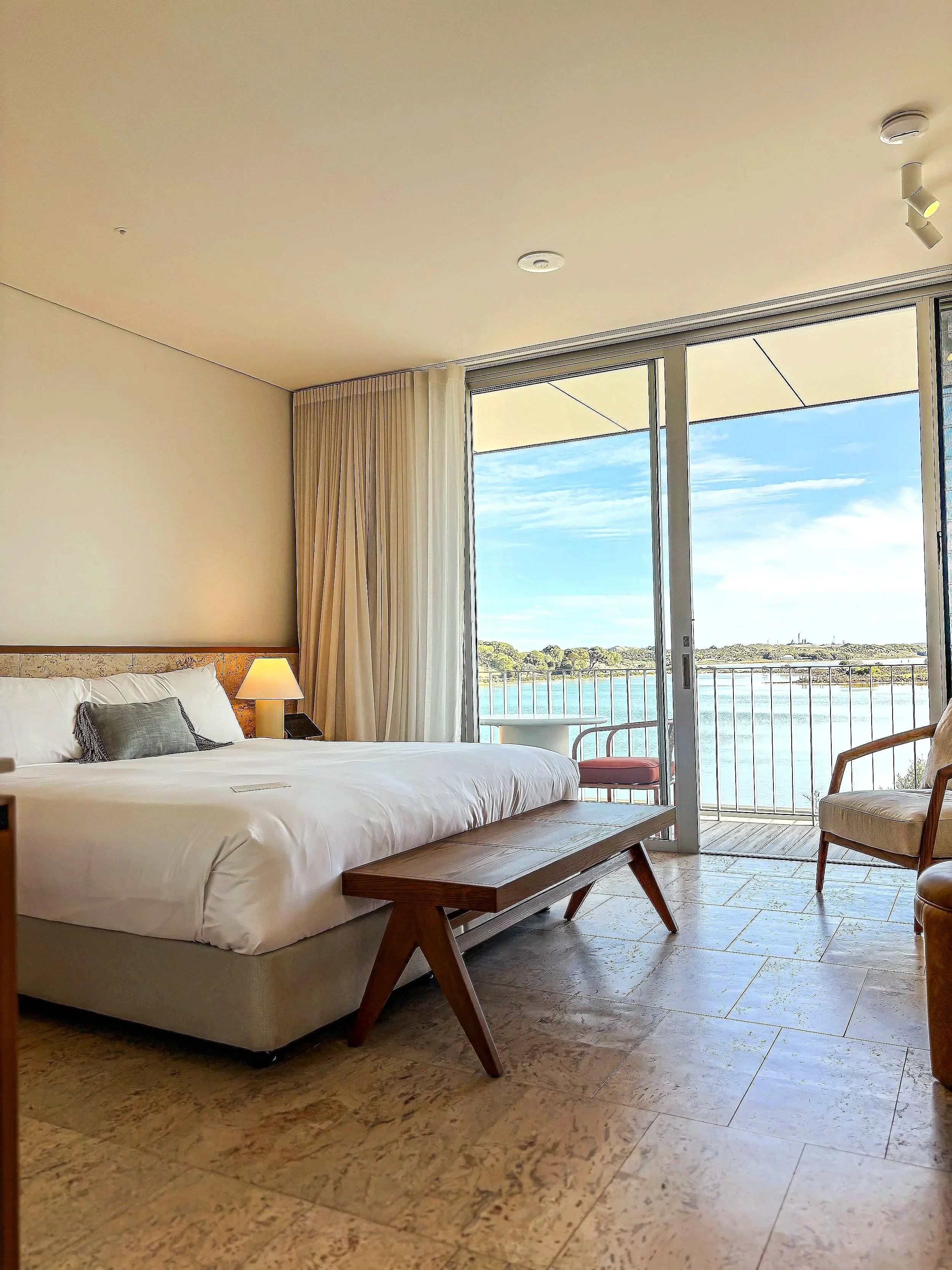 Hotel room with a large bed, bedside lamp, wooden bench at the foot of the bed, patio door leading to a balcony, water view outside, chairs on the balcony, beige curtains, and tiled floor.