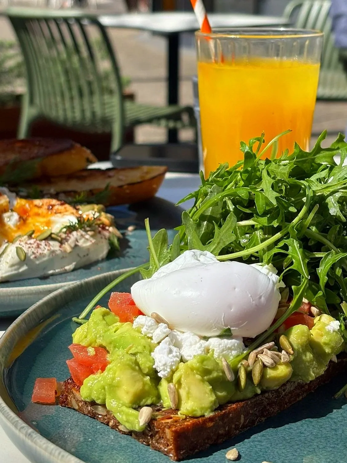 Close-up of avocado toast with poached egg, tomatoes, greens, sunflower seeds, served on a dark ceramic plate. In the background, a glass of orange juice with a striped straw, and part of a breakfast dish with bread, on a blue plate. Outdoor setting with chairs.
