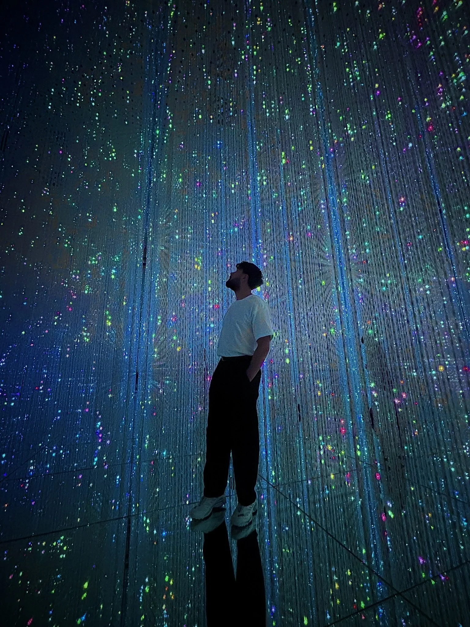 A person standing in an immersive art installation with hanging lights and holographic projections, creating a starry effect.