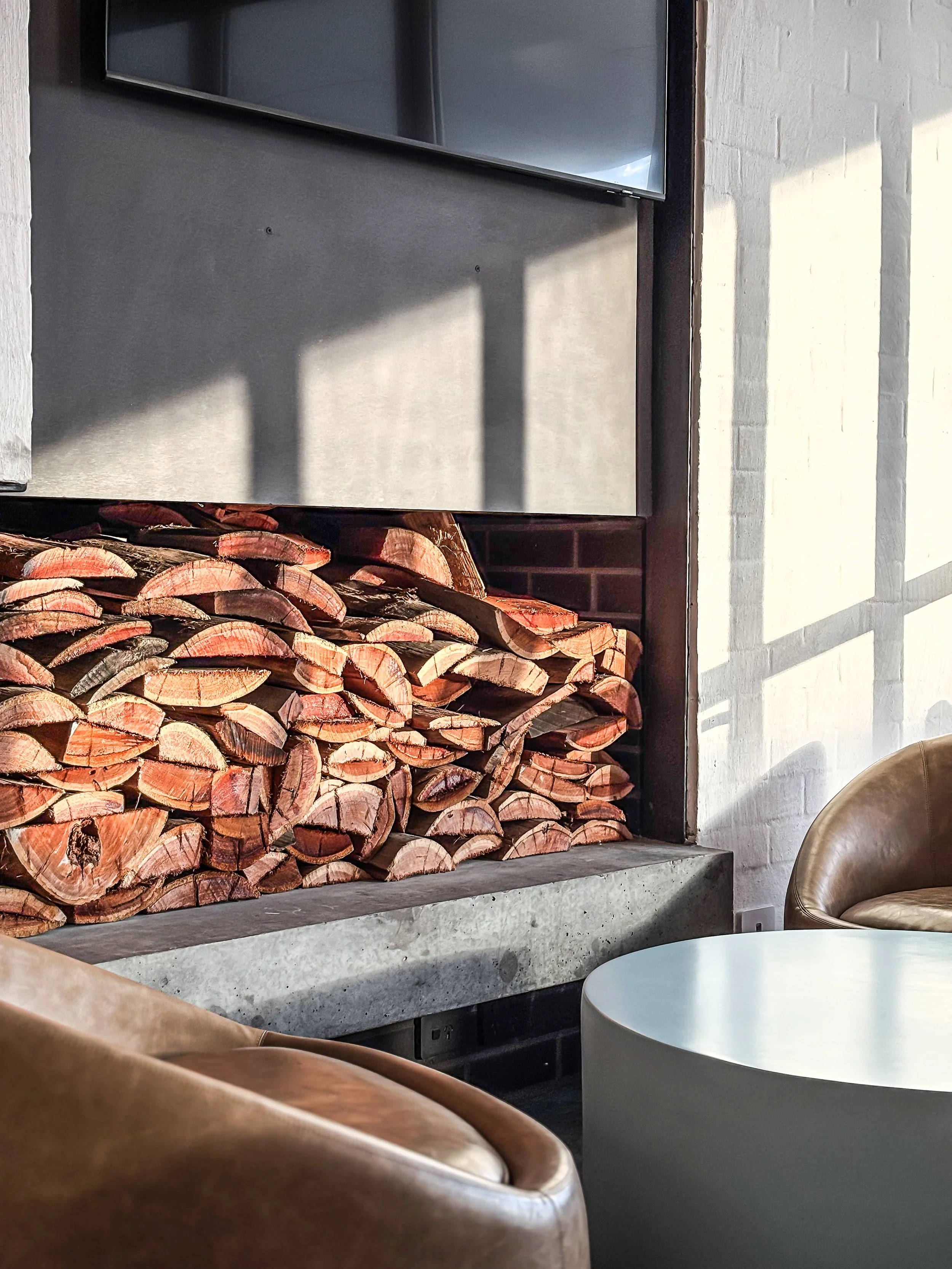 A cozy indoor space with a stack of firewood logs, a concrete hearth, modern furniture including a light brown leather chair, a white round table, and a wall with artificial lighting and shadows