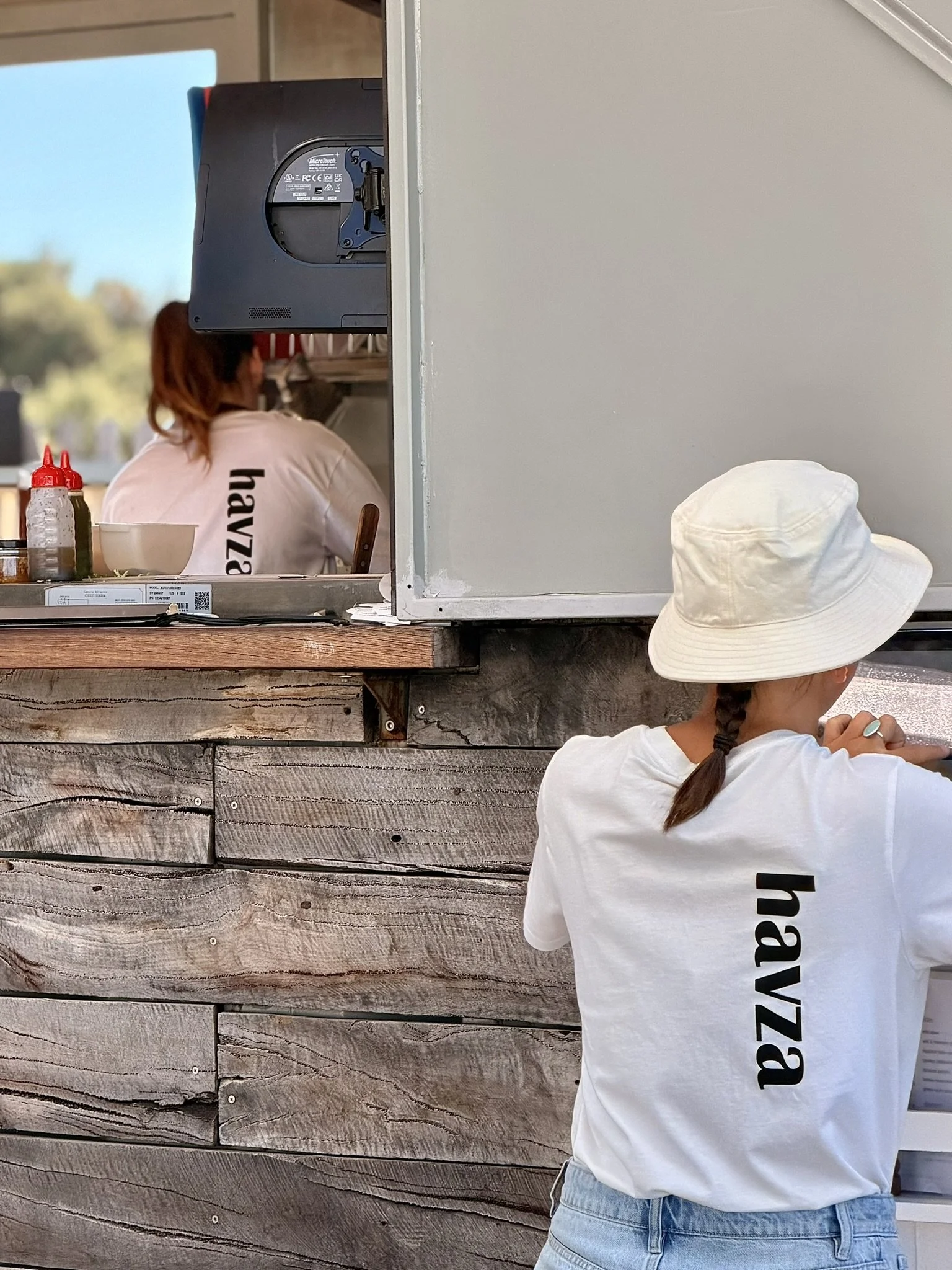 Person wearing a white hat and a white T-shirt with the word 'hazy' printed on the back, standing at a counter, talking to a person inside a food truck.