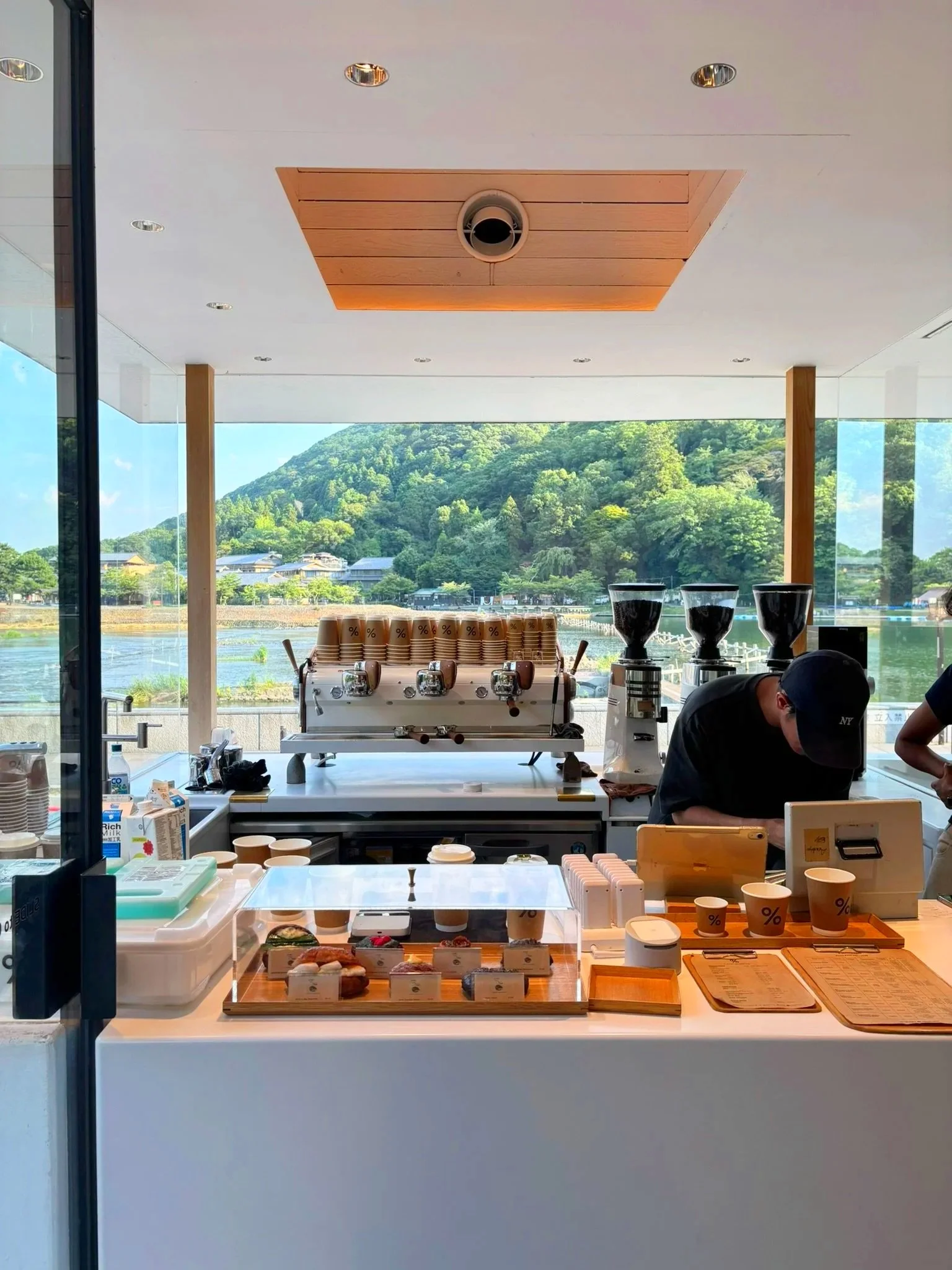 A coffee counter inside a cafe with an espresso machine, coffee cups, and desserts, overlooking a scenic view of a mountain and lake through large windows.