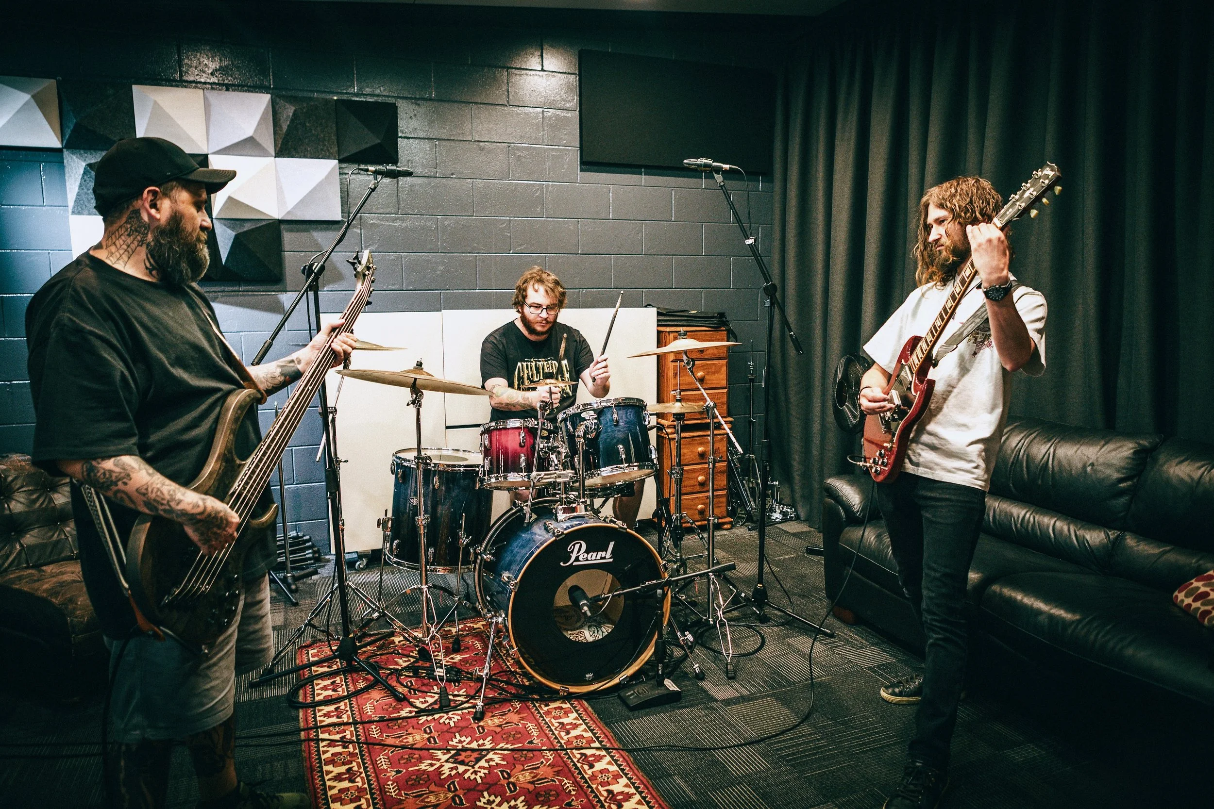 Three musicians practicing in a recording studio, with one playing bass guitar, one on drums, and one playing electric guitar.
