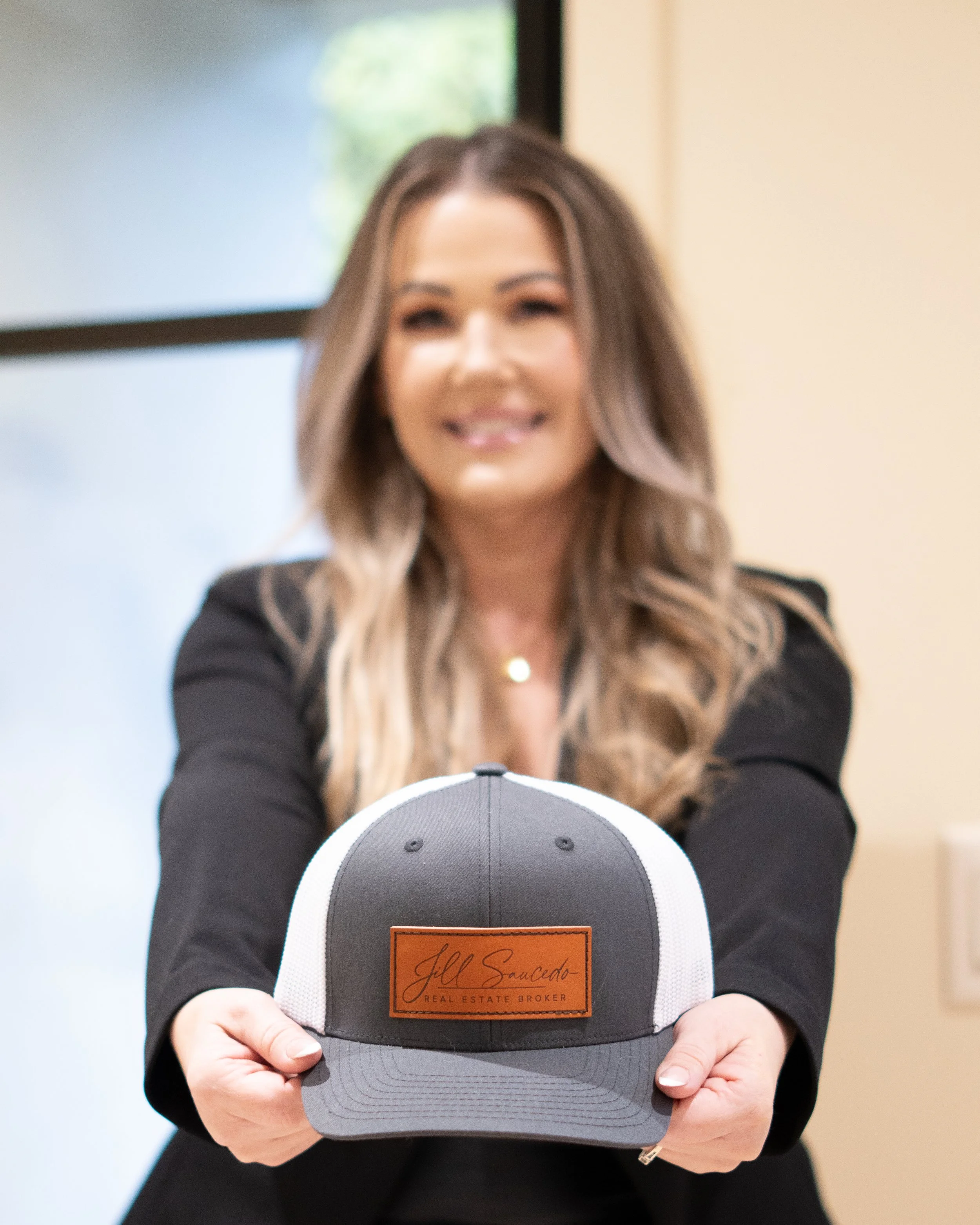 Portland metro real estate agent, A woman with long wavy hair smiling and offering a gray and white trucker cap with a leather patch reading "Jill Saucedo, Real Estate Broker" towards the camera.