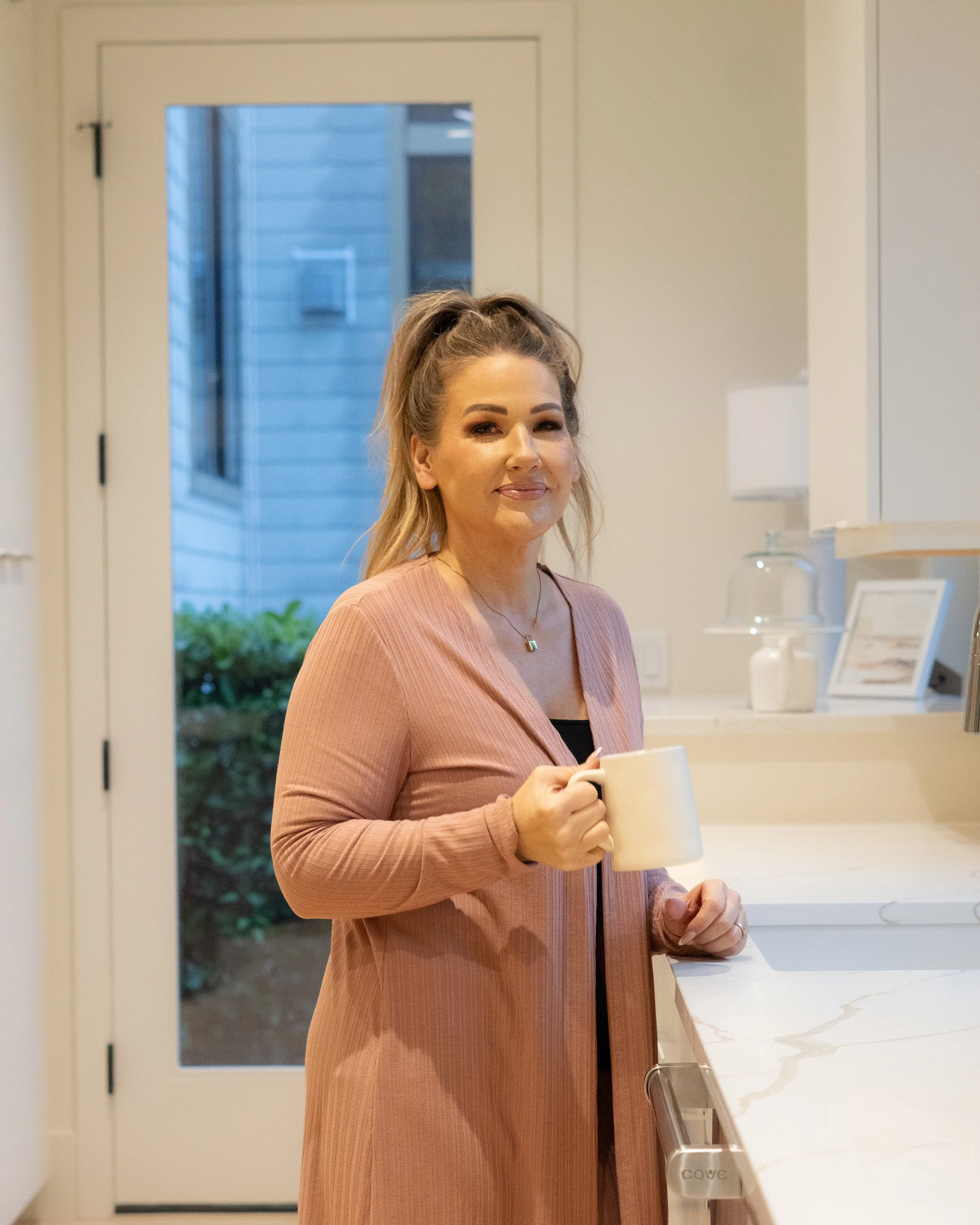 Jill Saucedo, Portland metro real estate agent, with blonde hair in a high ponytail, wearing a pink robe, standing in a kitchen holding a beige mug, smiling at the camera.