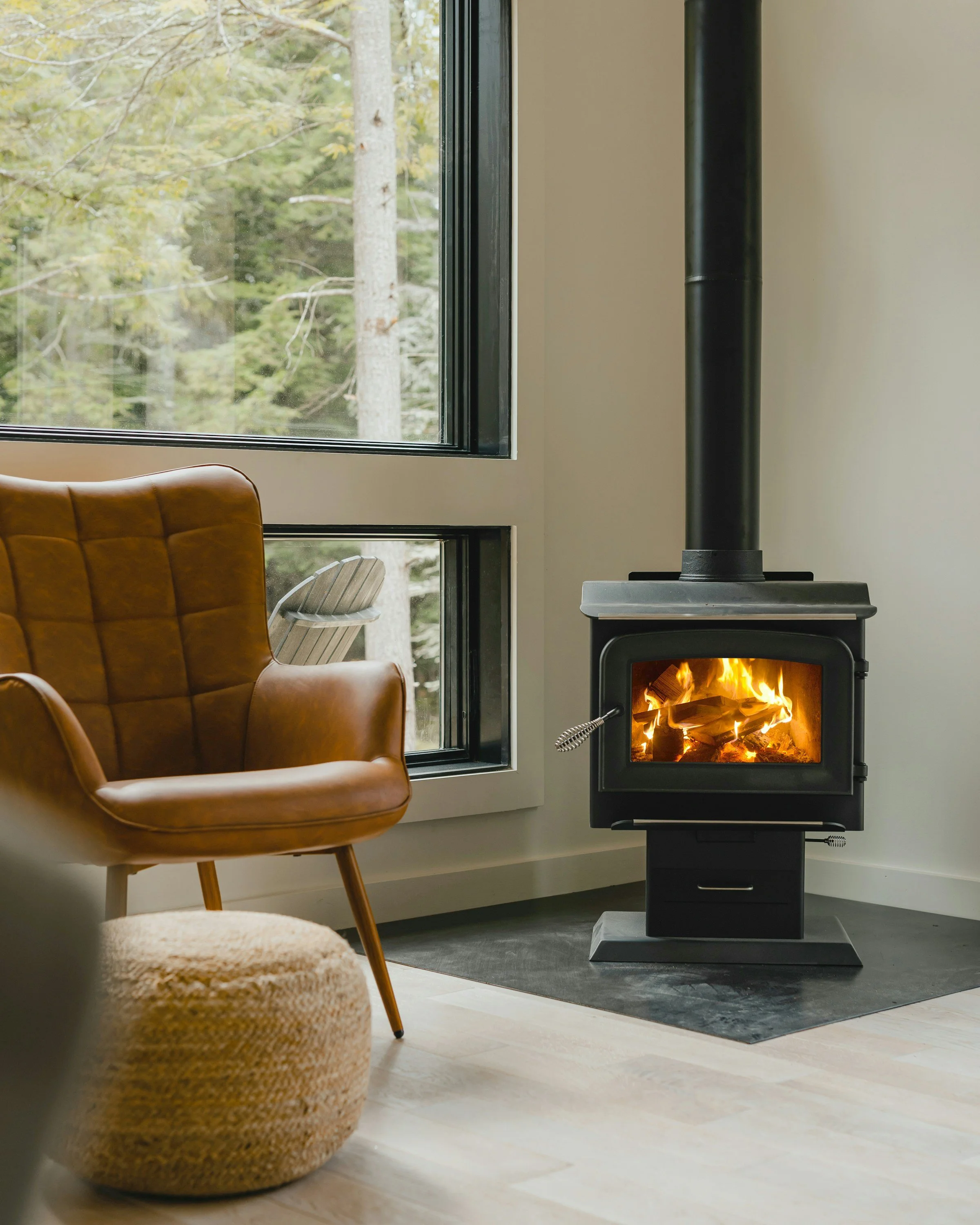 Portland metro real estate agent, Interior of a cozy room with a wood stove casting a glow, a tan leather armchair, and large windows showing a forested landscape outside.