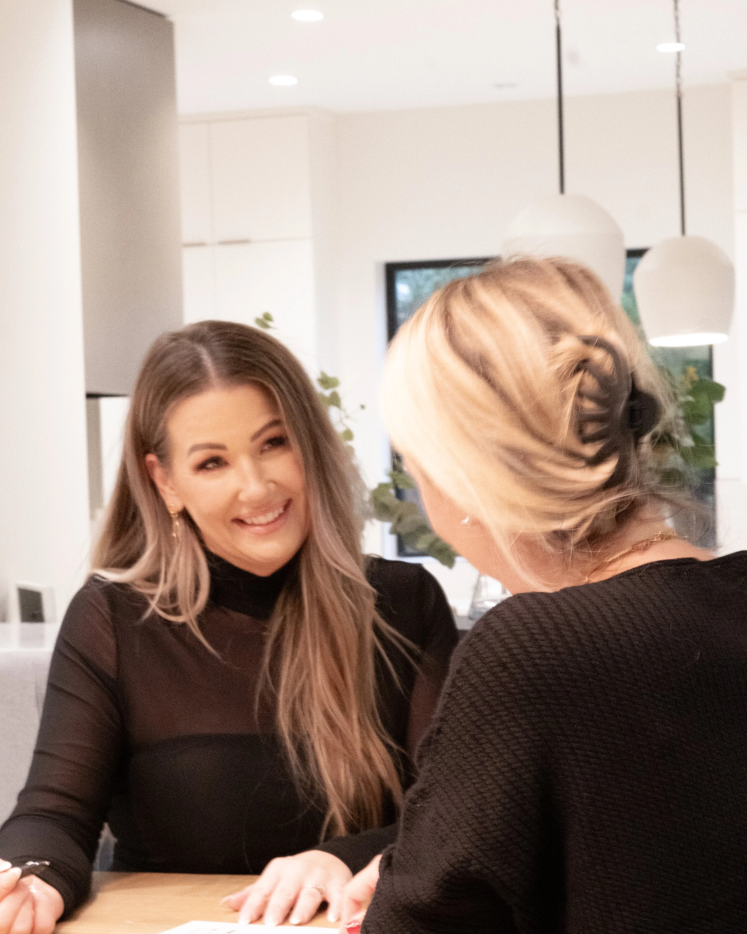 Portland metro real estate agent, Two women sitting at a table engaged in conversation indoors, with a modern home interior in the background.