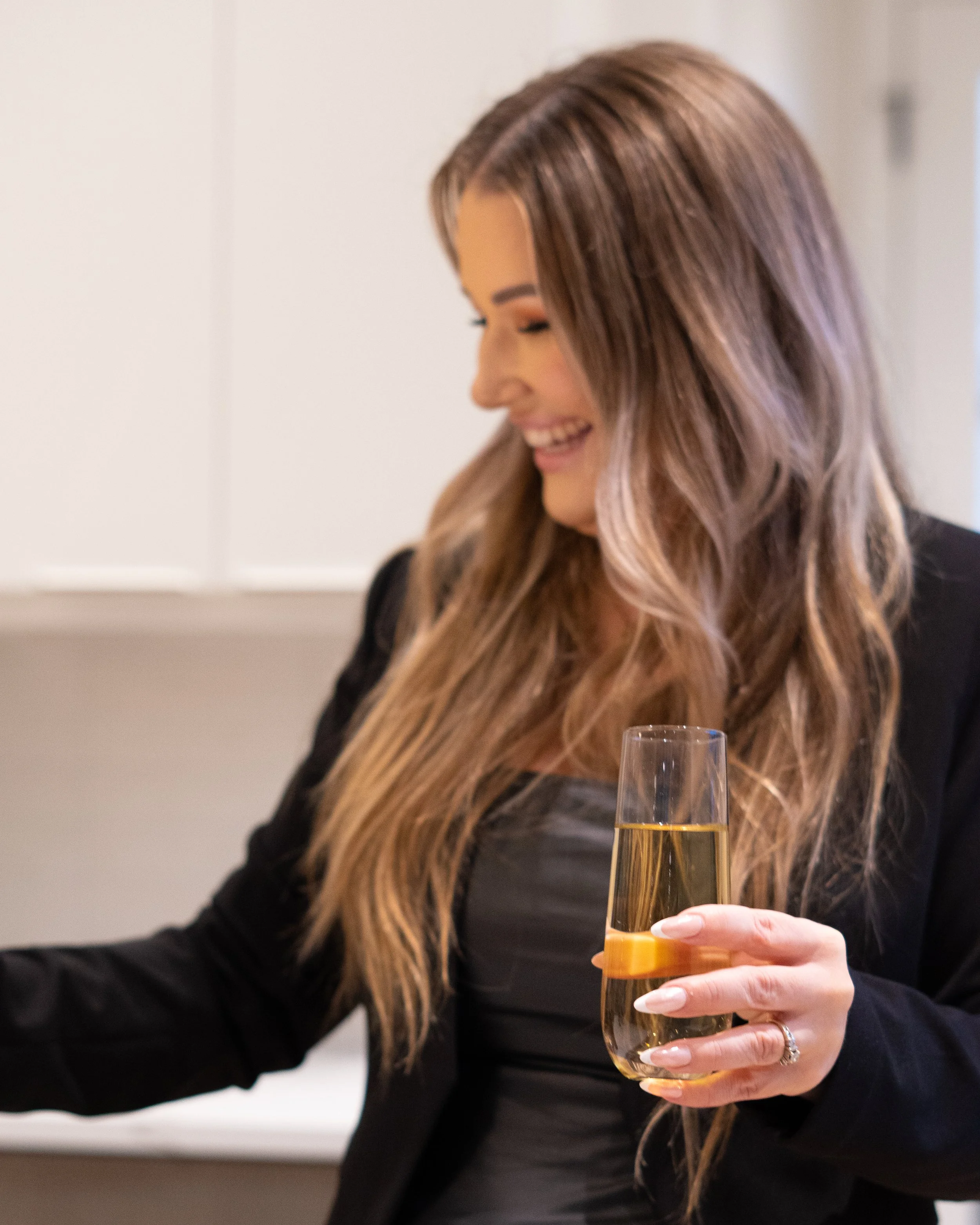 Jill Saucedo, Portland metro real estate agent, with long brown hair in a black blazer holding a glass of champagne, smiling and looking down.