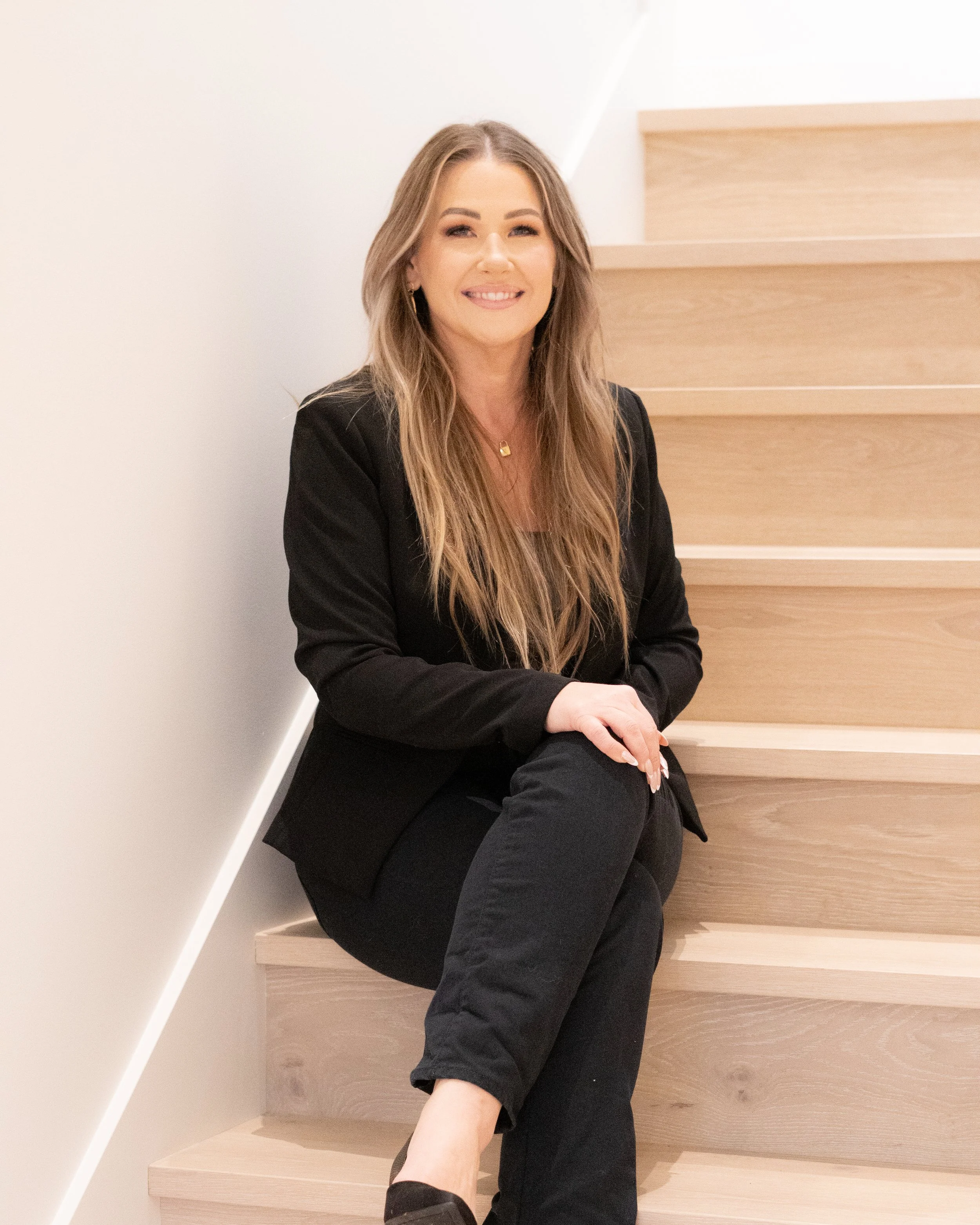 Jill Saucedo, Portland metro real estate agent, with long hair, dressed in black, sitting on wooden stairs, smiling at the camera.