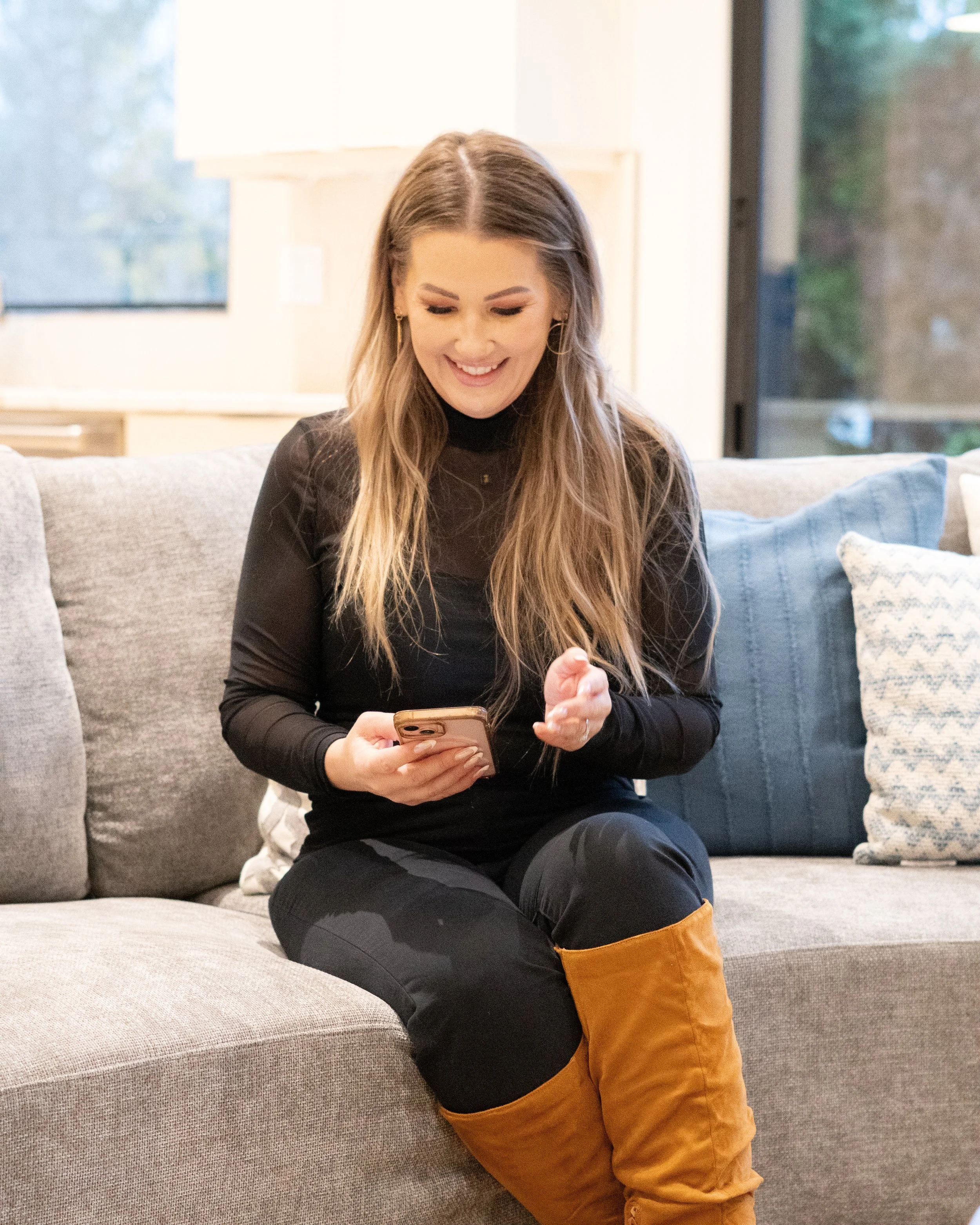 Jill Saucedo, Portland metro real estate agent, with long hair, wearing a black outfit and tan boots, sitting on a light gray sofa and looking at her phone in a living room.