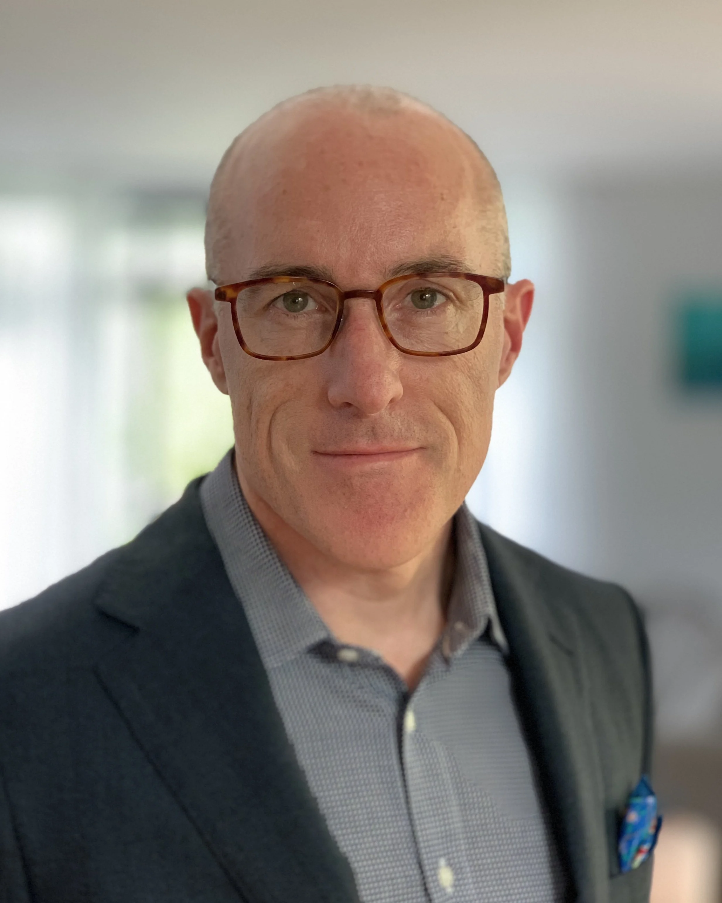 Portrait of Lindsay O'Sullivan, Founder of Tenet Advisory, wearing glsses, a gray shirt and a dark blazer, smiling in a well-lit indoor setting.